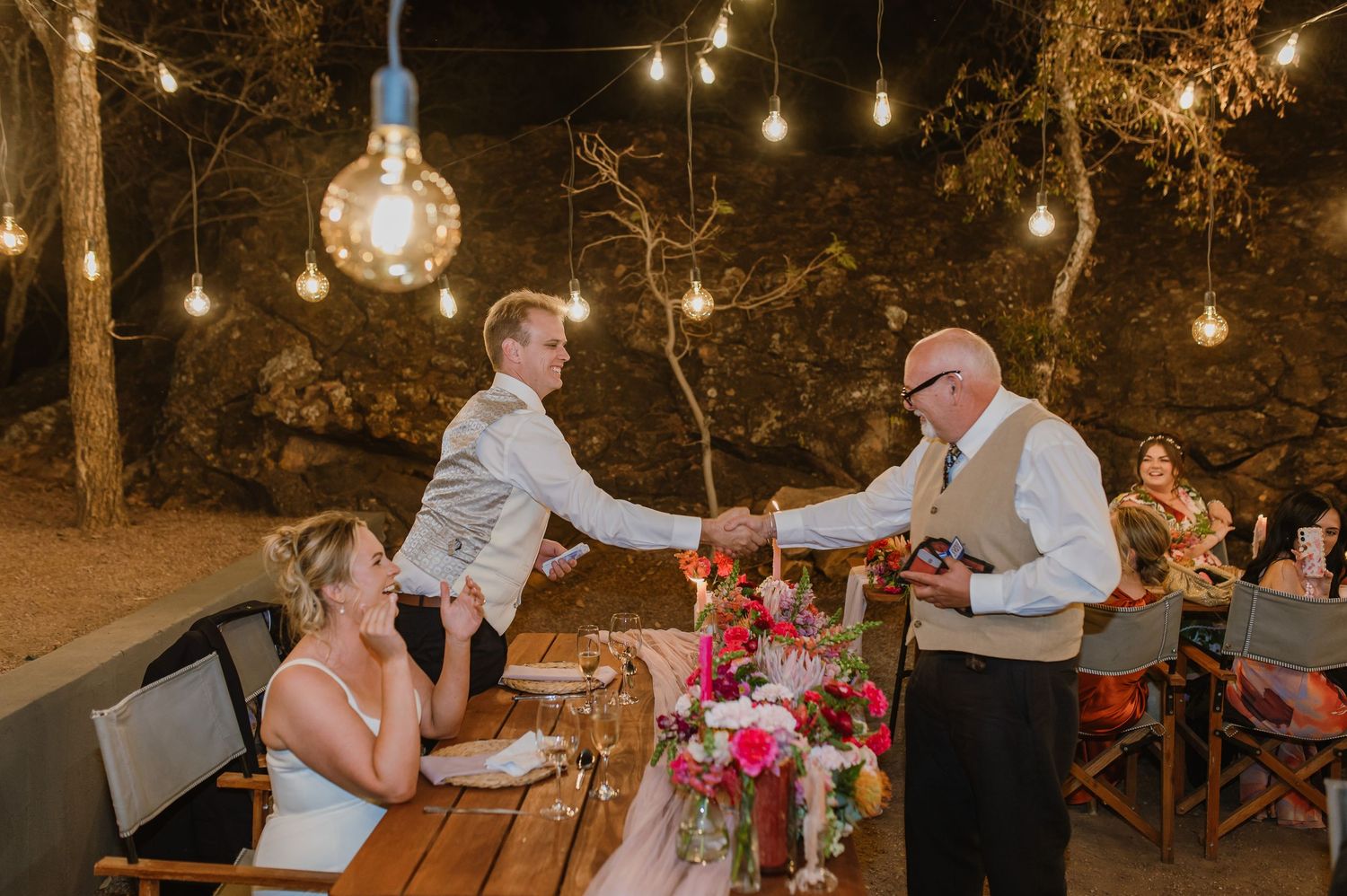 Guests enjoy an intimate outdoor dinner under string lights at dusk with rustic wooden tables and floral centerpieces.