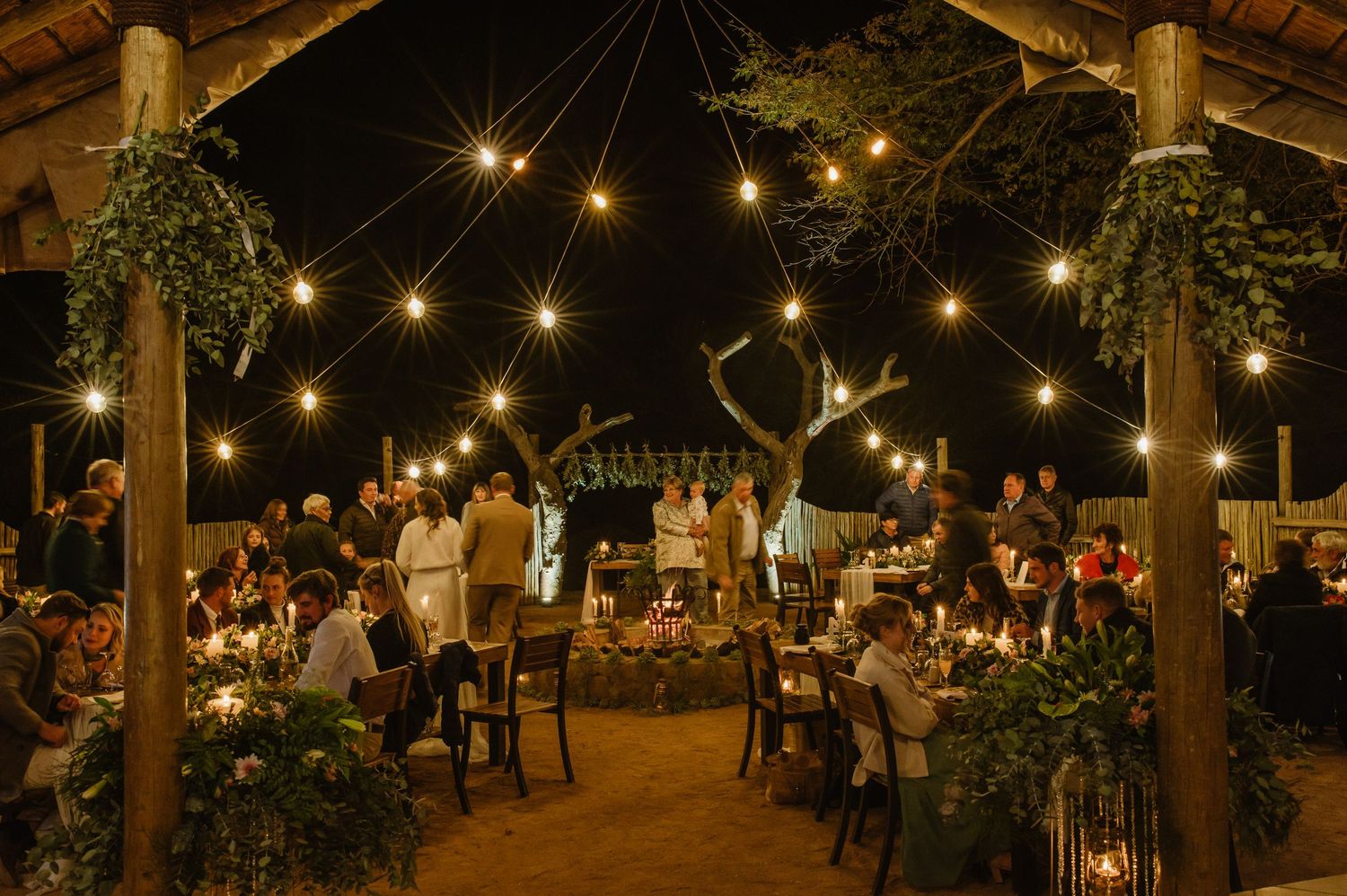 String lights illuminate an outdoor evening reception area with rustic wooden tables and chairs.