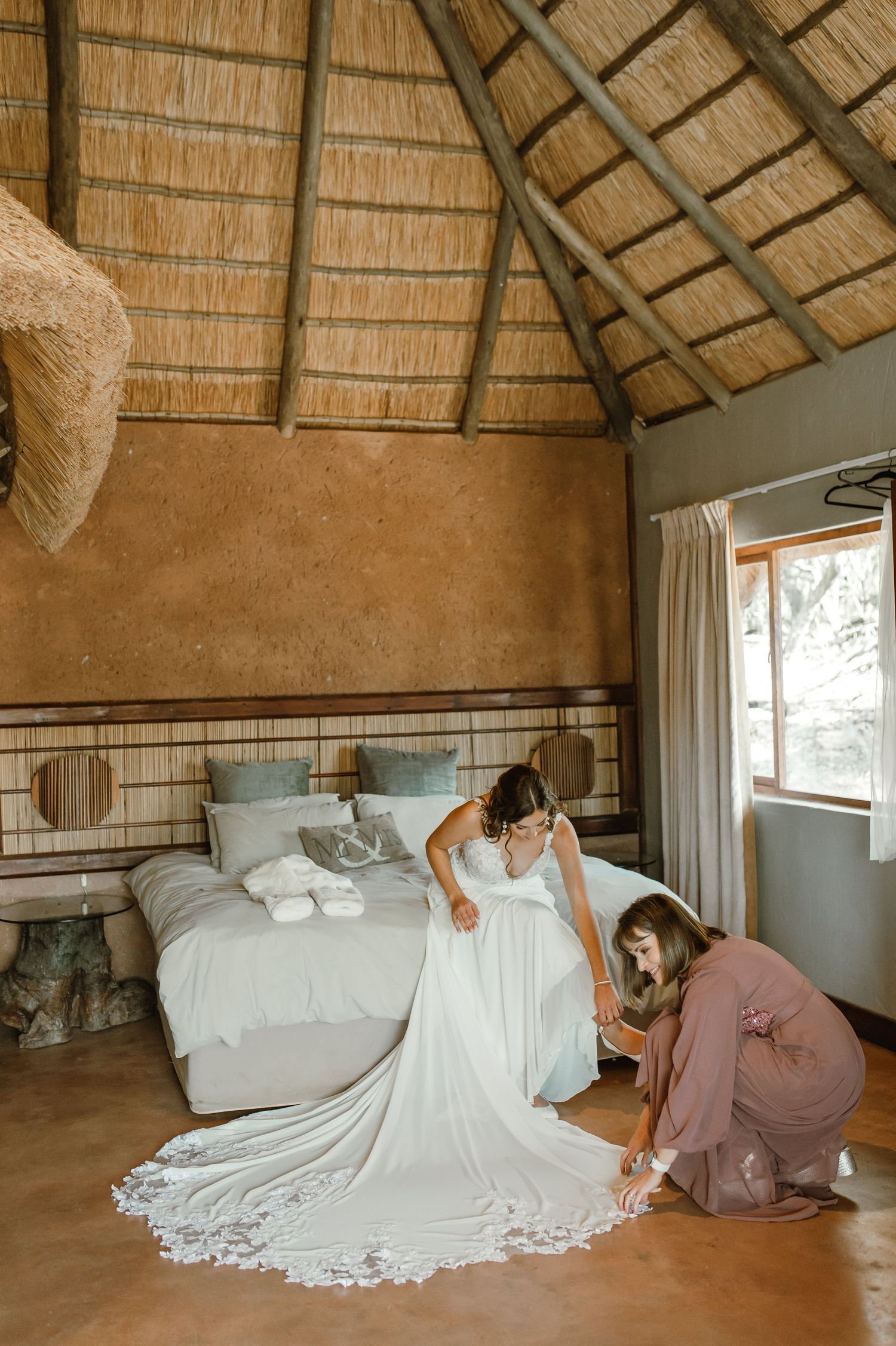 White wedding dress being arranged on a bed in a rustic room with thatched roof.