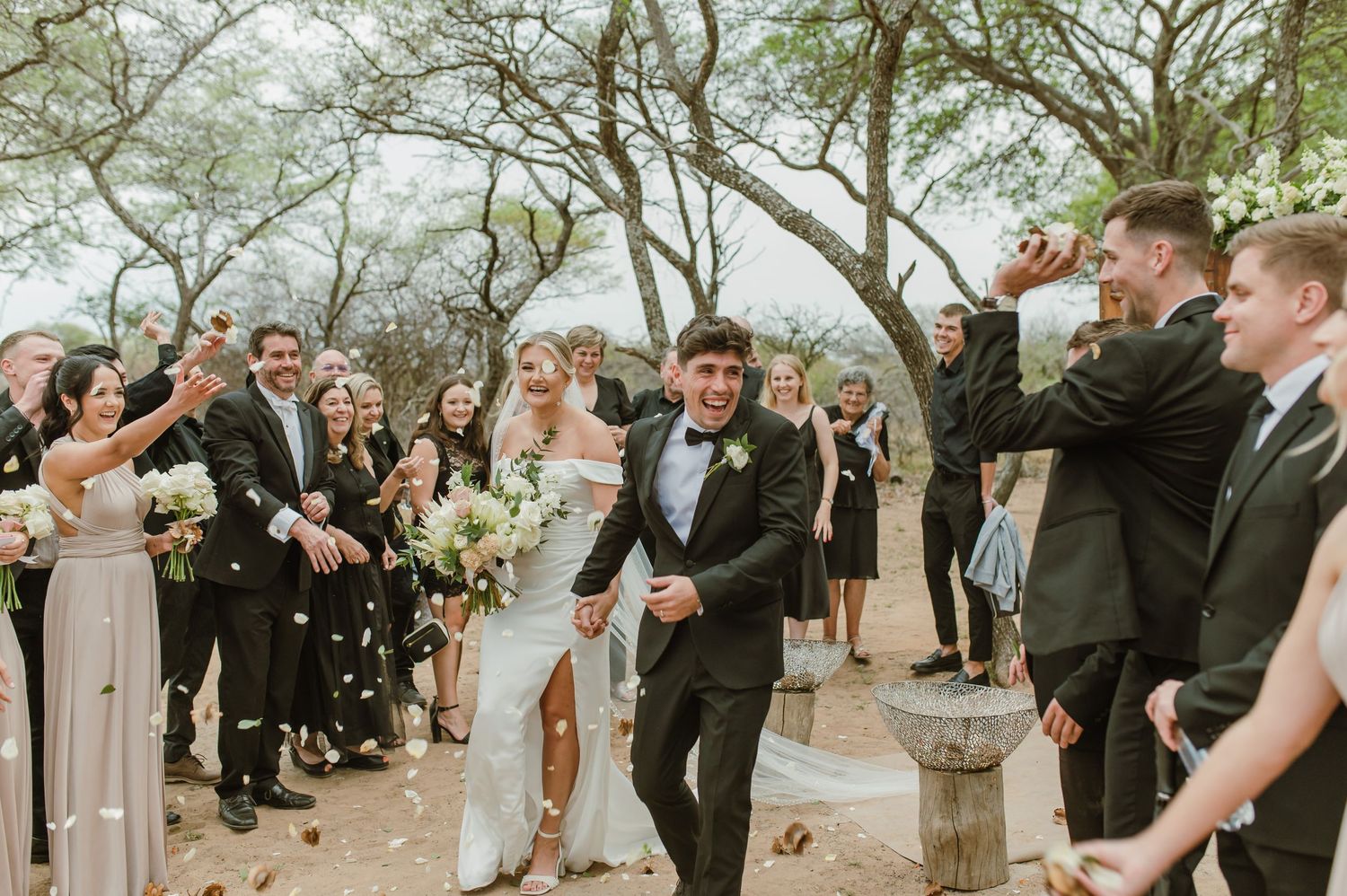 Wedding party celebrates with confetti toss on a tree-lined path.