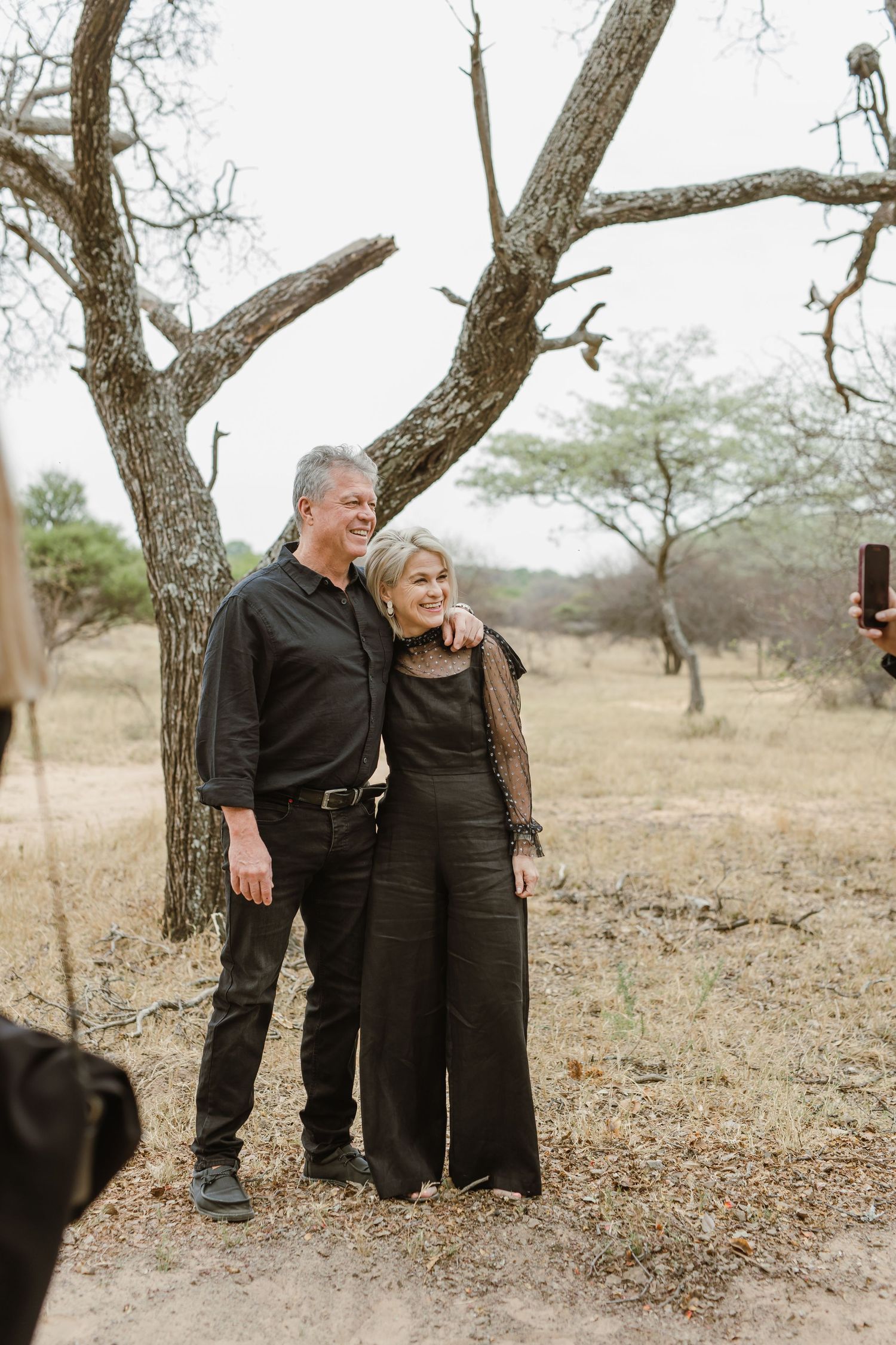 Couple in black formal wear share an intimate moment under a tree in a natural setting.