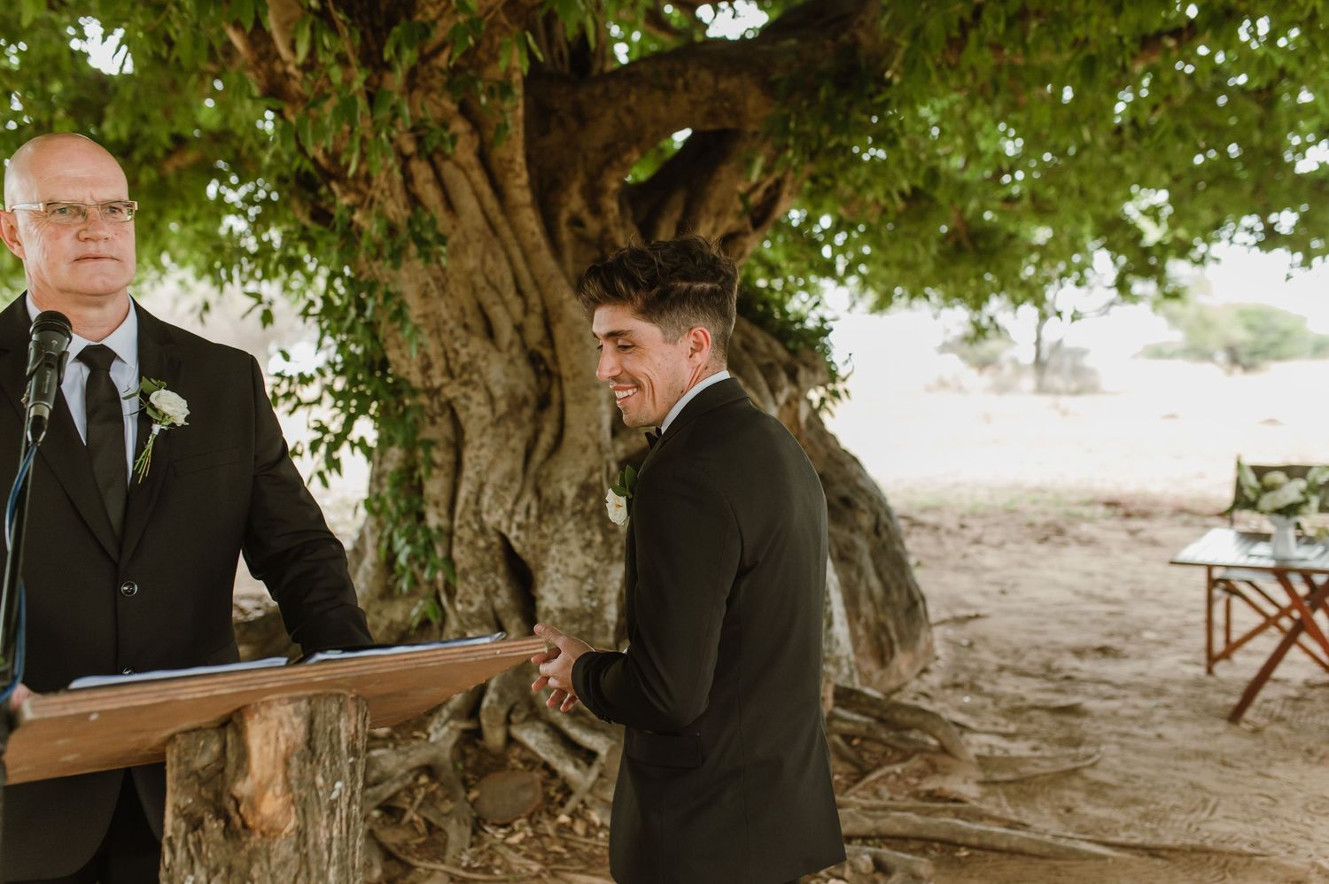 Wedding ceremony guests gather under a large tree canopy during an outdoor celebration.