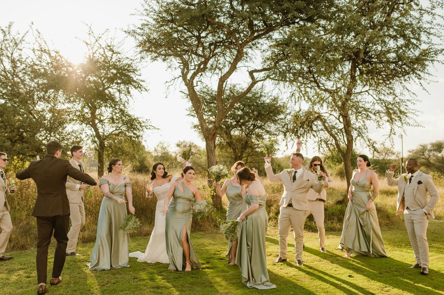 Wedding party celebrates outdoors at sunset with sage green dresses and tan suits under acacia trees.