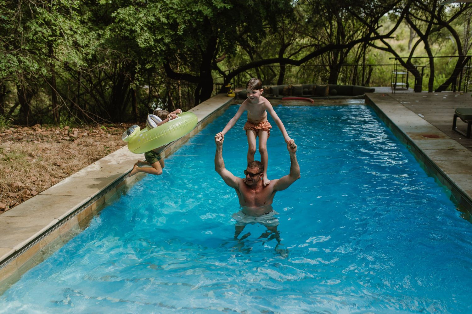 Father lifting child above water in a serene outdoor swimming pool surrounded by trees.