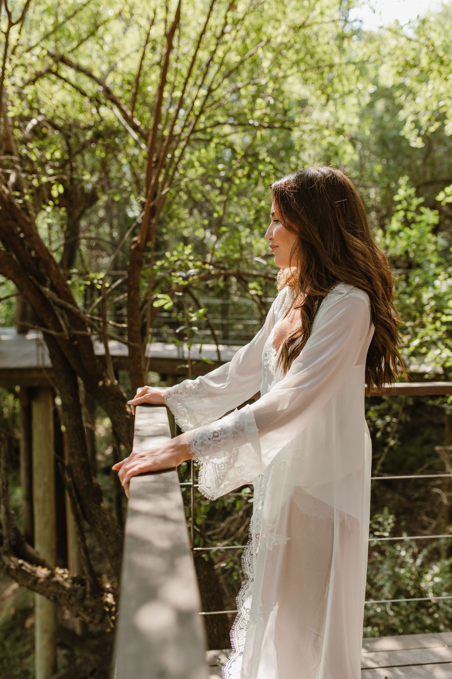Bride in white lace robe standing on wooden deck balcony surrounded by green trees on wedding day.