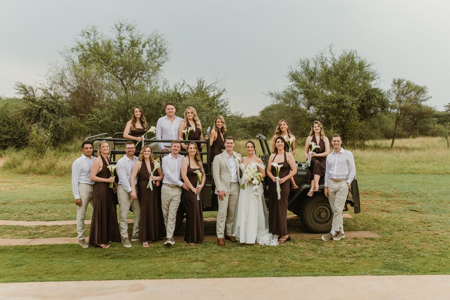 A wedding party poses together on a grassy lawn with trees in the background on a sunny day.