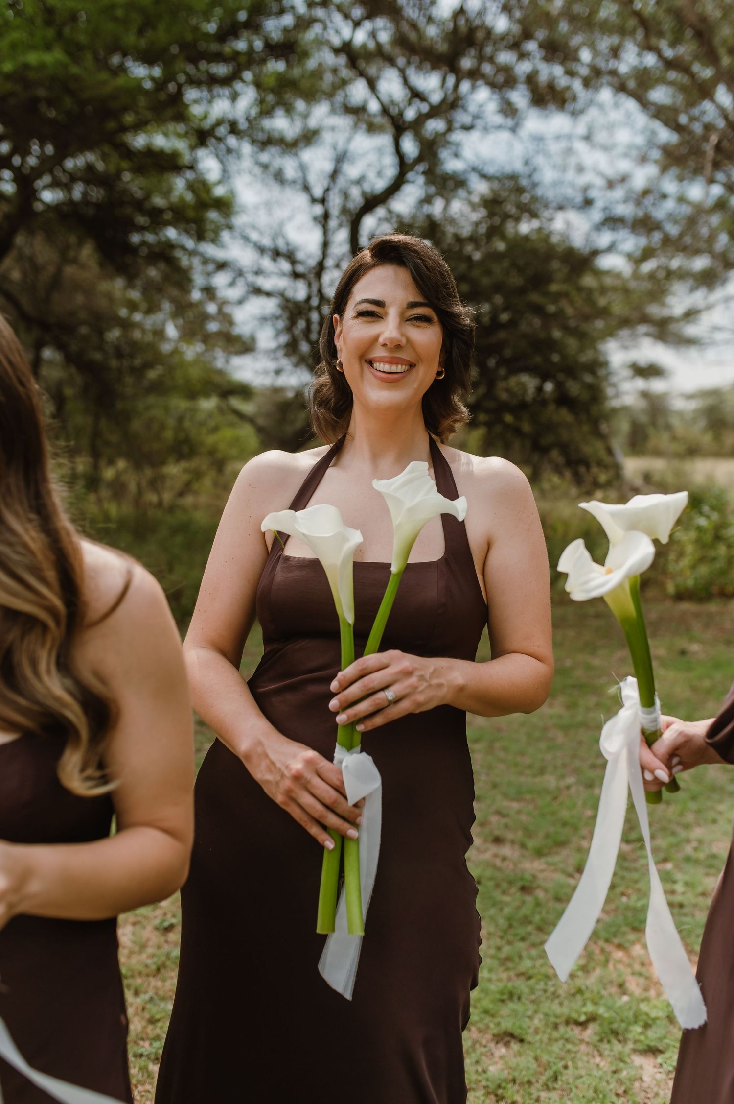 Wedding party members in brown dresses hold white calla lilies during an outdoor photo session.