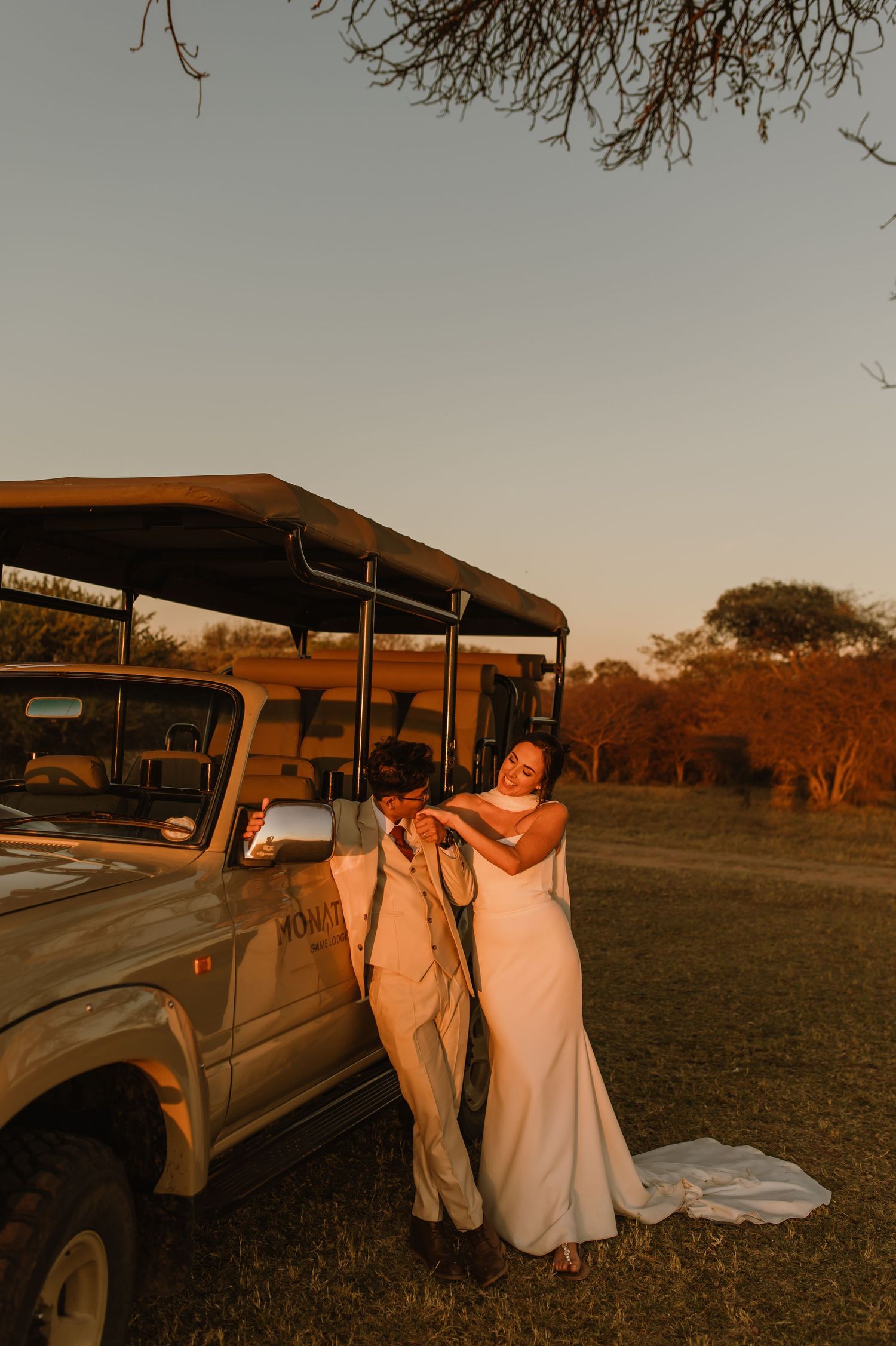Bride and groom share a romantic moment beside a safari vehicle at golden hour in an African bush setting.