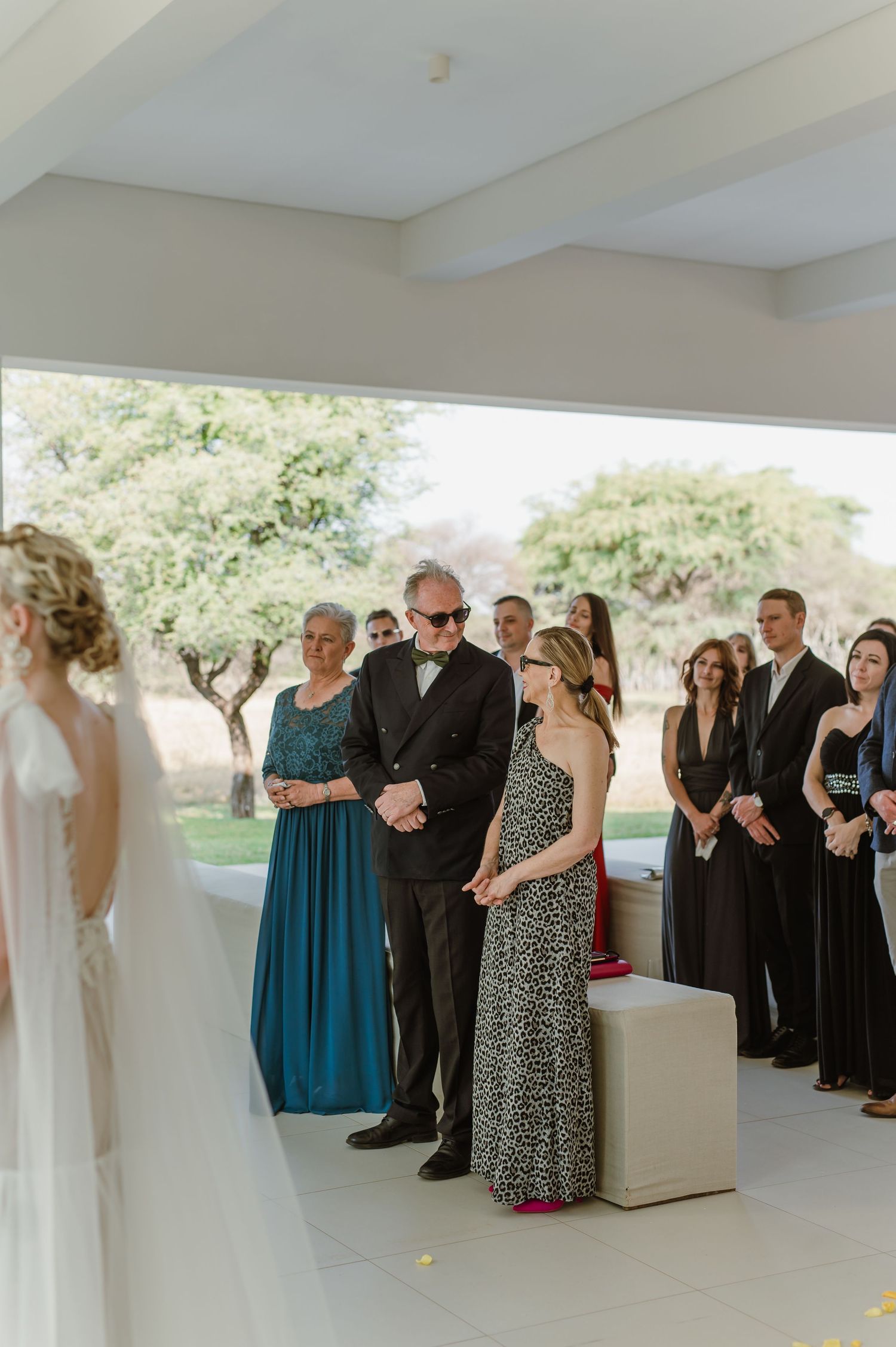 Wedding attendees in formal attire stand in a modern white space with large windows.
