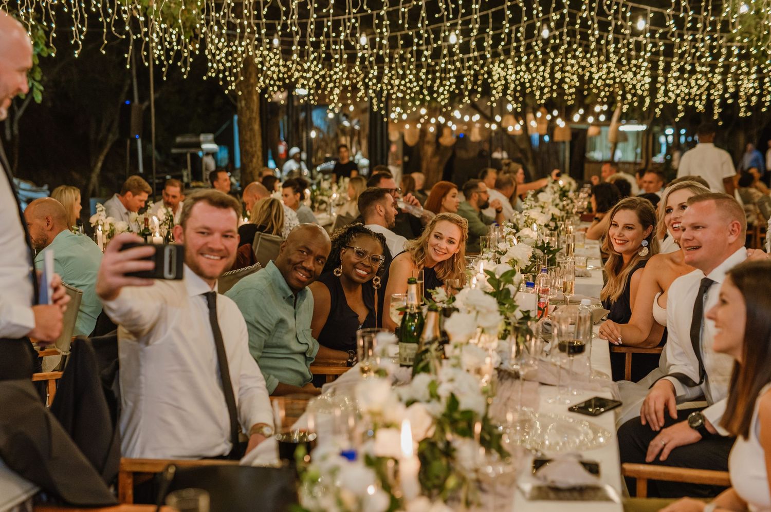 Wedding reception venue decorated with hanging lights and white floral centerpieces.