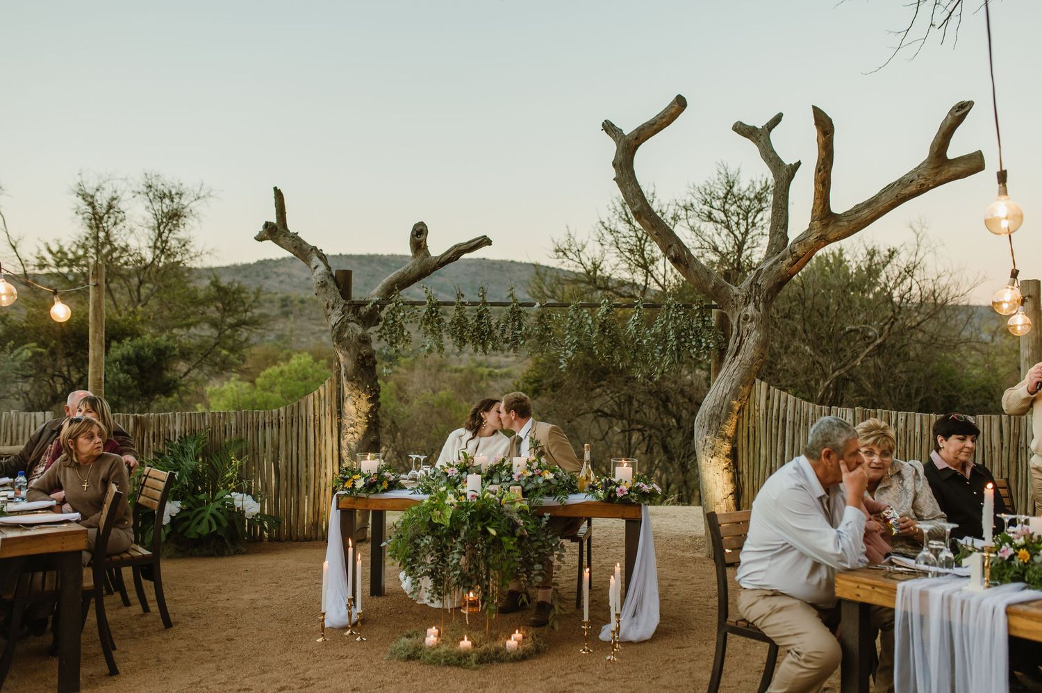 Rustic outdoor wedding reception venue at sunset with string lights and wooden tables set for dining.