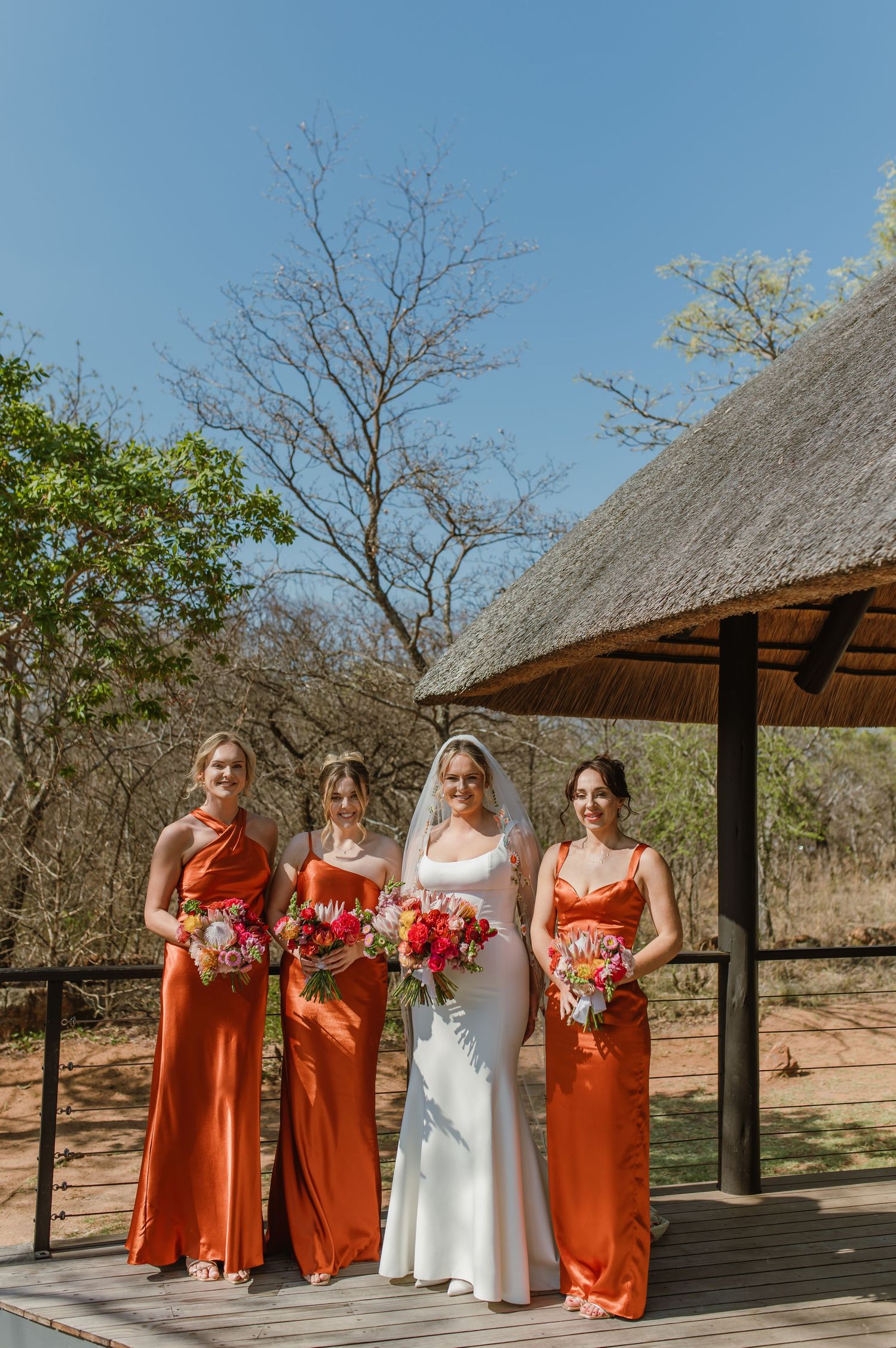 Wedding party in orange dresses poses under a thatched roof pavilion in a safari setting.