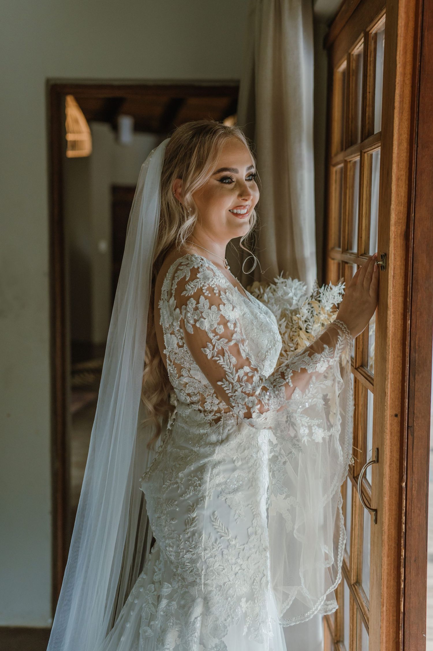 Bride in long-sleeve lace wedding gown poses elegantly by wooden window frames.