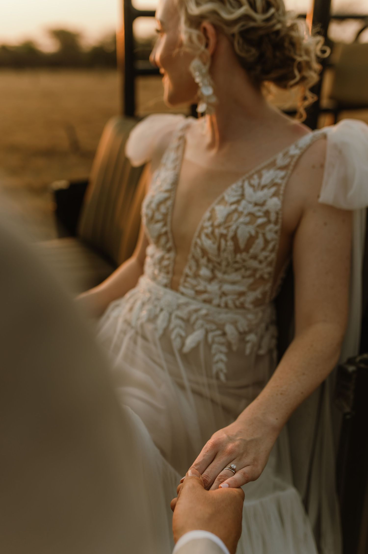 Close-up detail of a bride's lace wedding dress and clasped hands in warm sunset light.