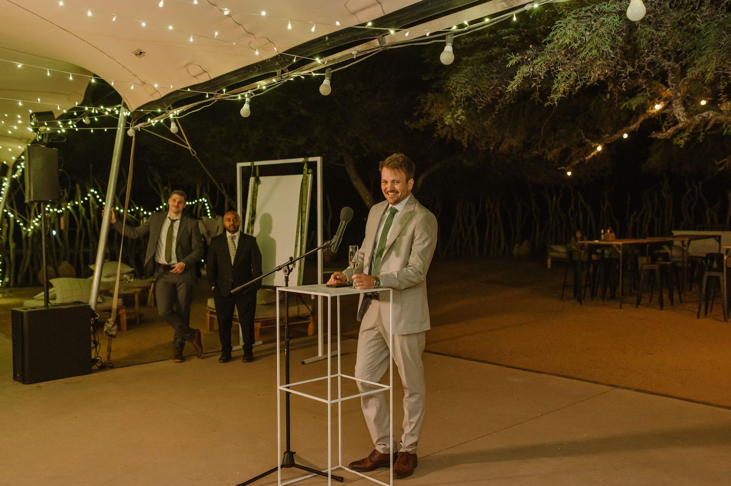 Evening wedding reception speech under a tent with string lights and greenery.