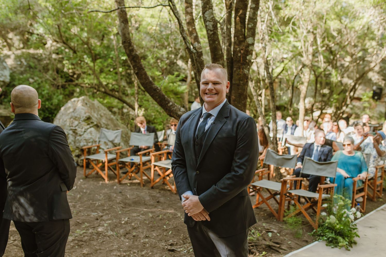 Guests seated in wooden chairs observe a ceremony in a wooded outdoor setting.