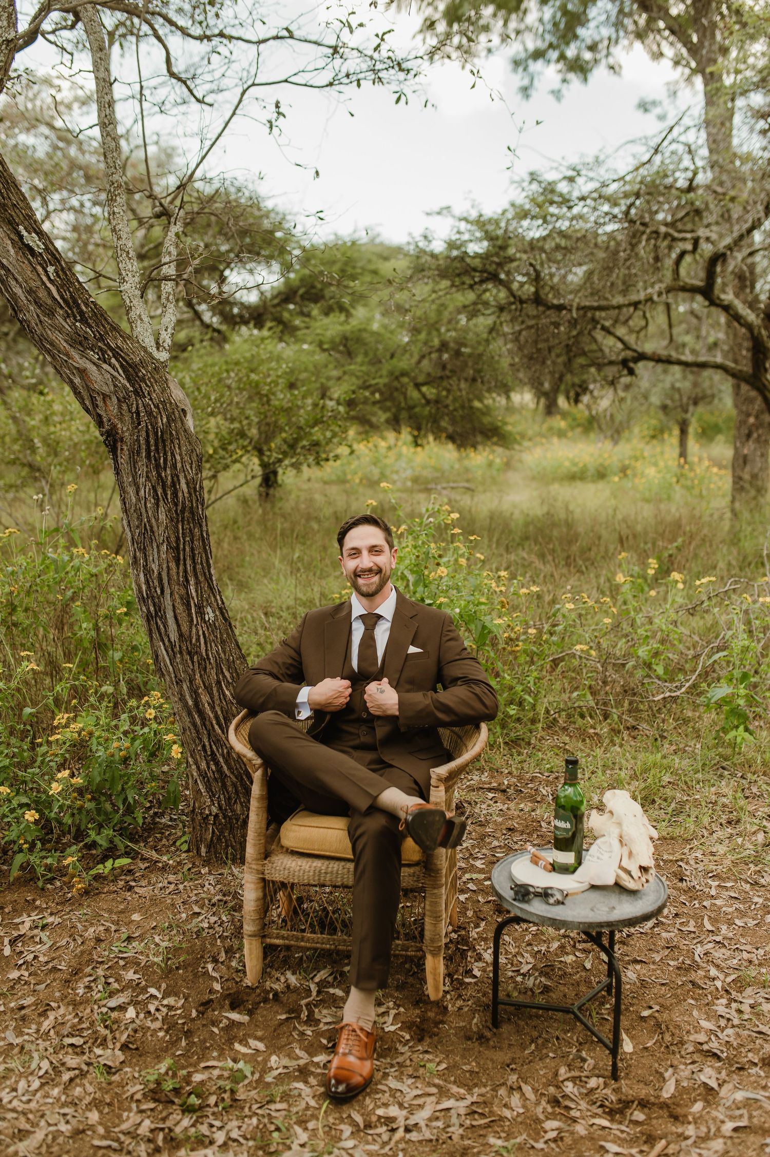 Person in brown suit sitting in wicker chair with small table and refreshments in outdoor setting.