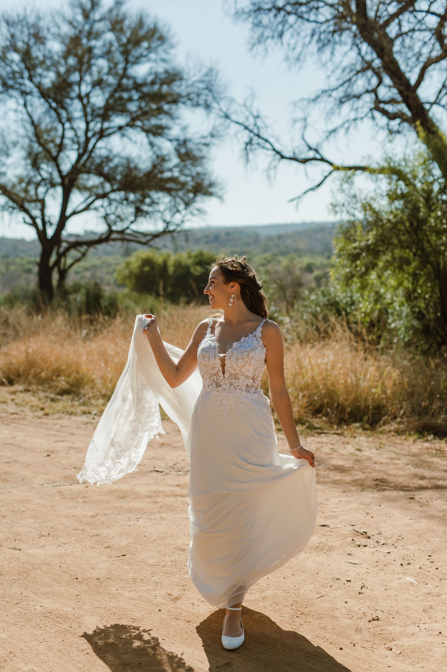 Bride twirls in flowing white wedding dress on desert path with mountains behind.
