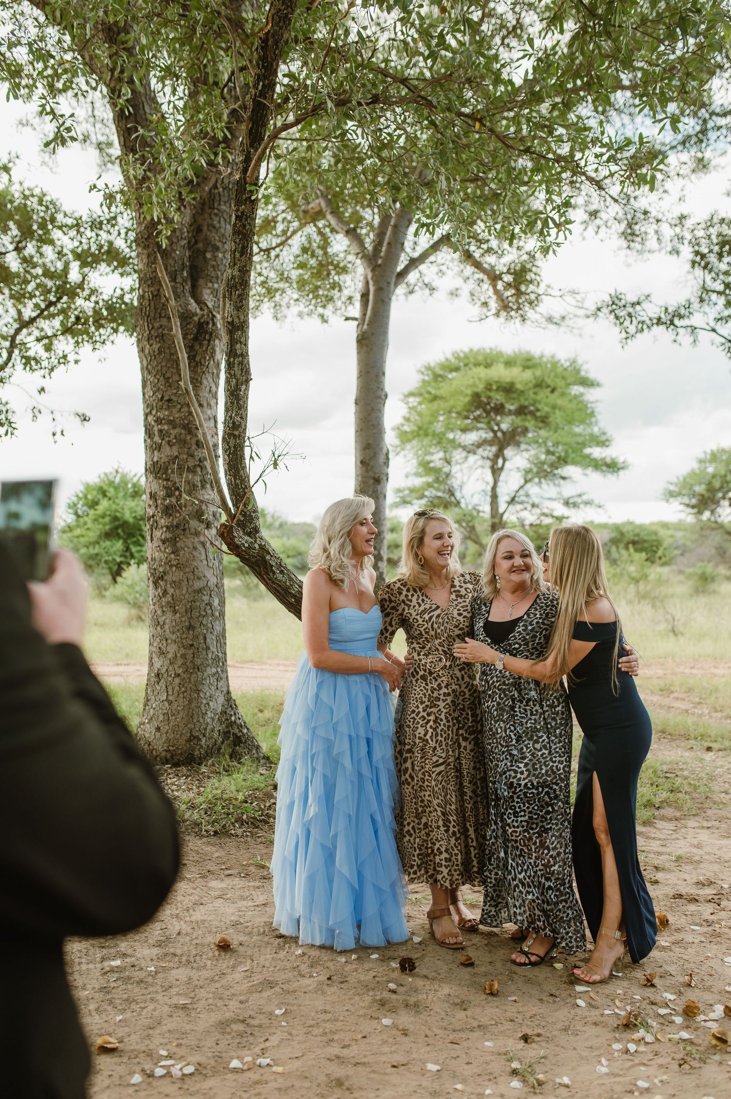 Wedding guests pose for photos under shaded tree grove.