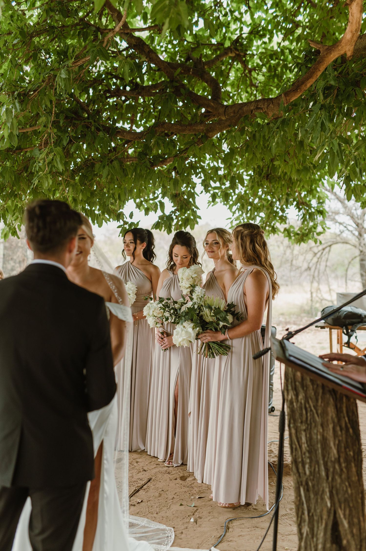 Bridesmaids in light colored dresses standing during outdoor wedding ceremony.