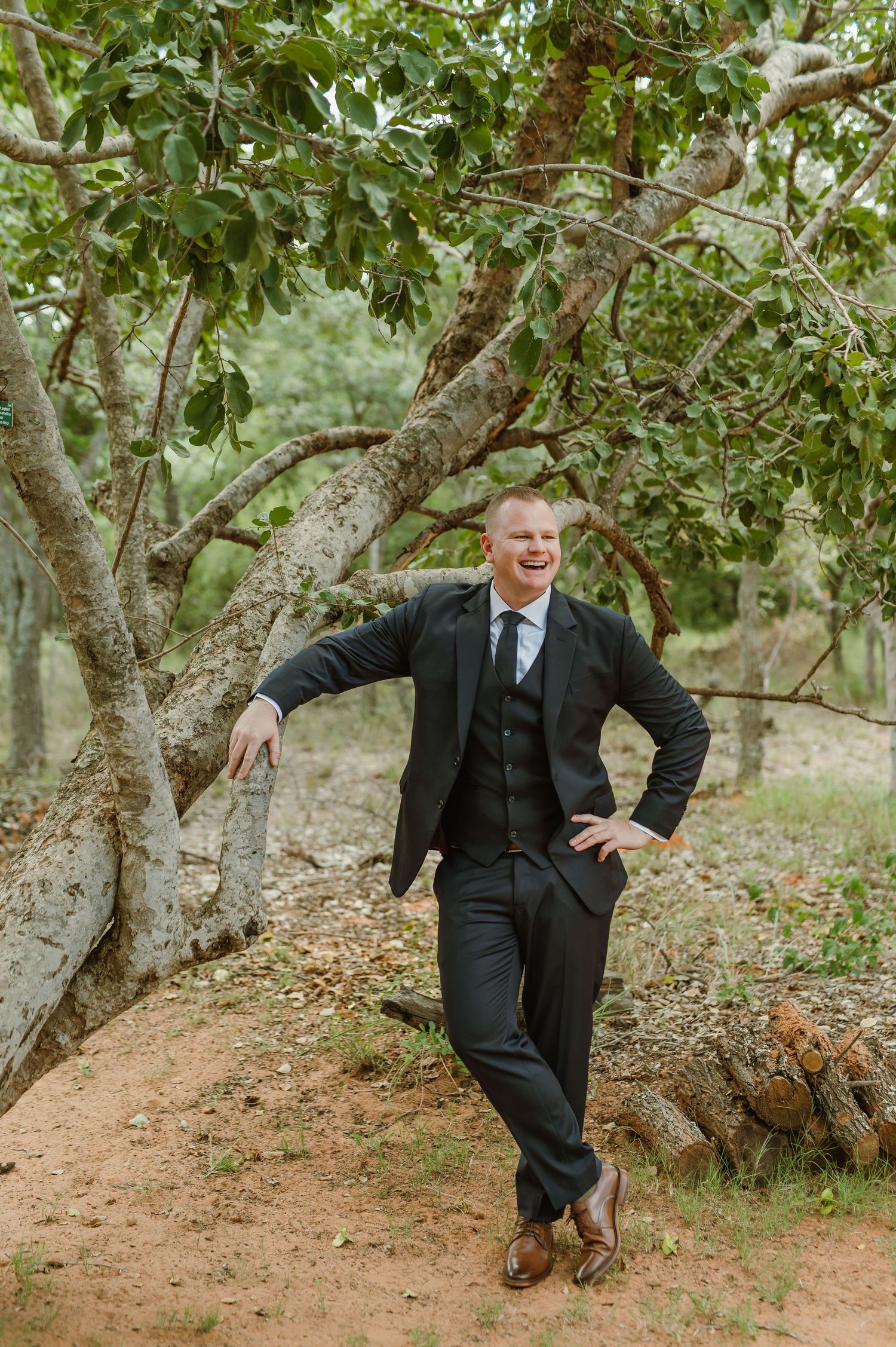 Groomsmen posing playfully by a tree in a rustic outdoor setting wearing black suits.