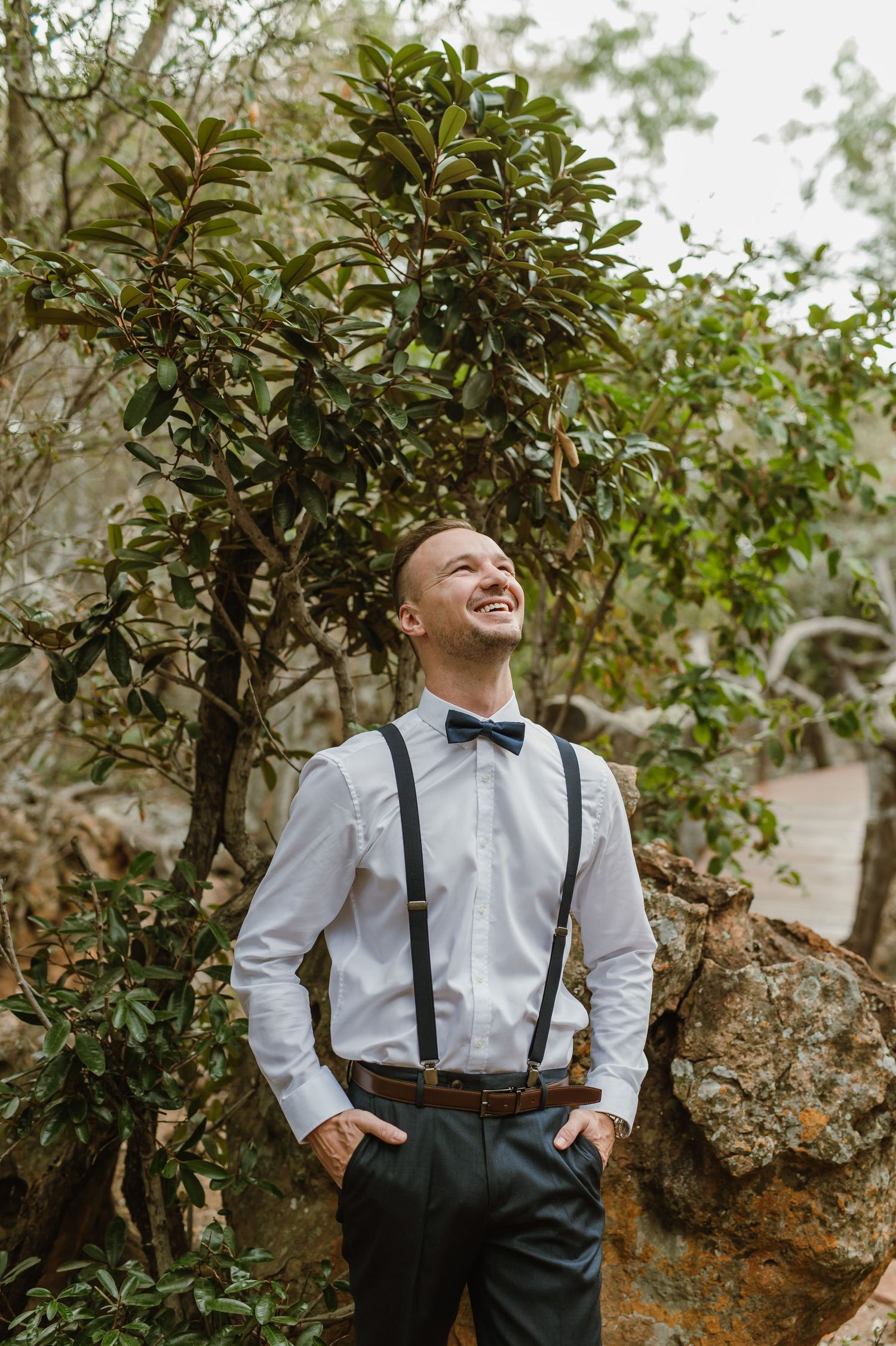 A groom in suspenders and bow tie poses casually against a tree in an outdoor setting.