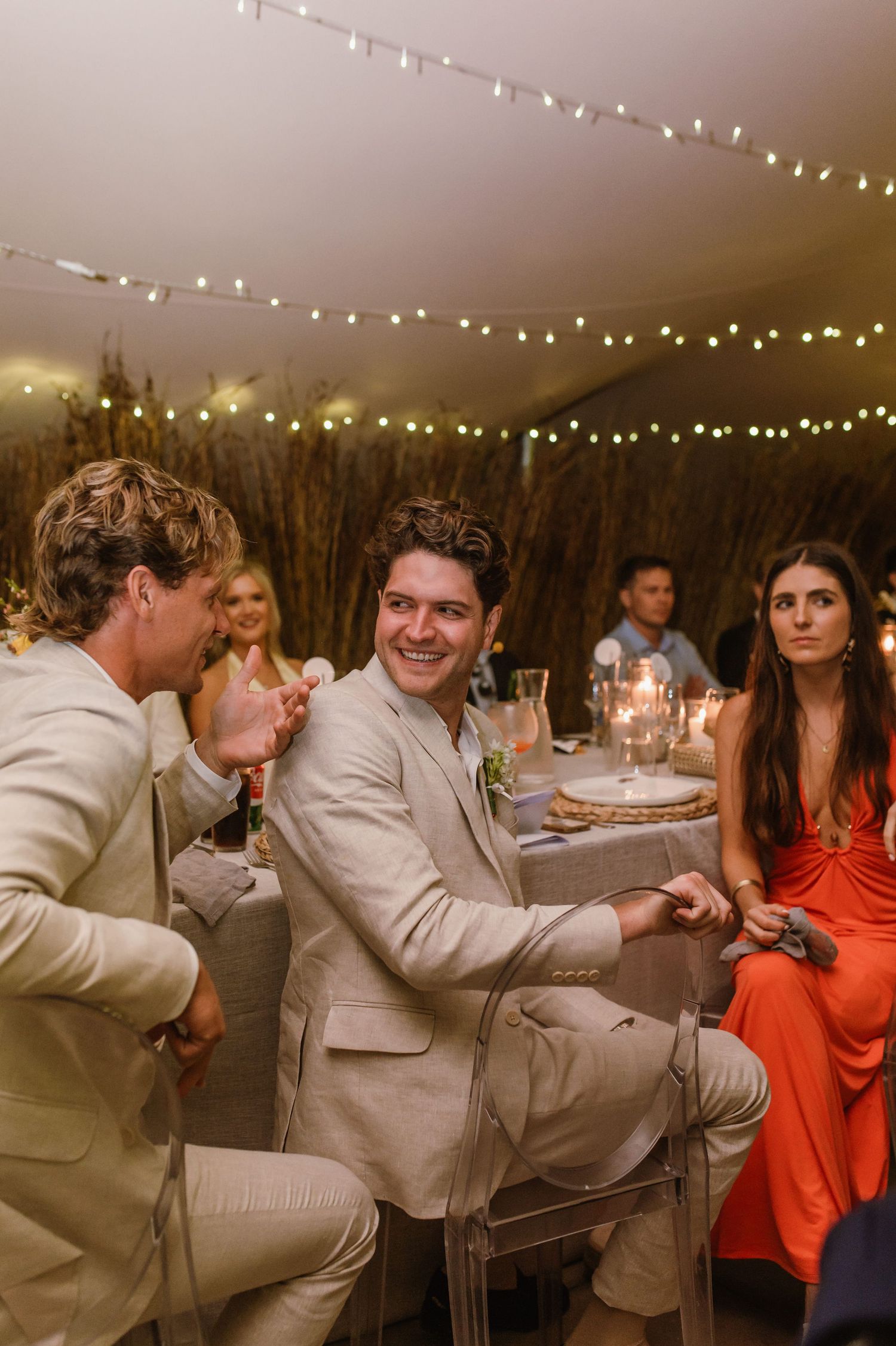 Wedding guests in formal attire socialize at tables under twinkling reception lights.