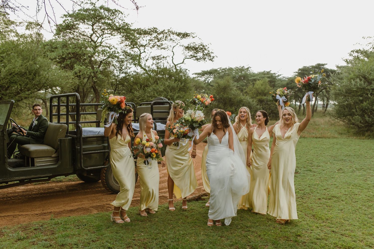 Wedding party walks together on dirt path carrying colorful bouquets with vintage truck in background.