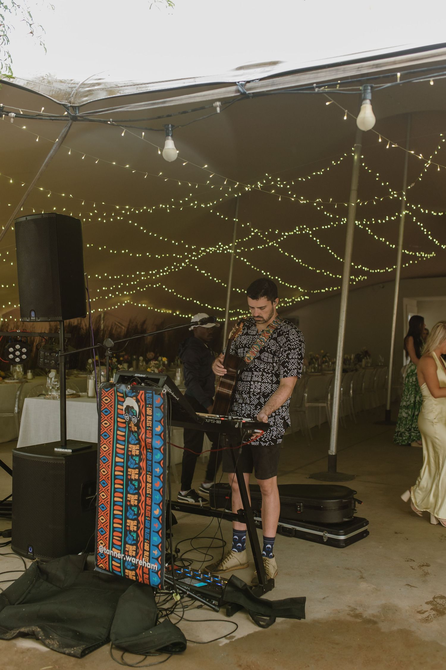 Musicians perform under string lights at a wedding reception with instruments and equipment set up.