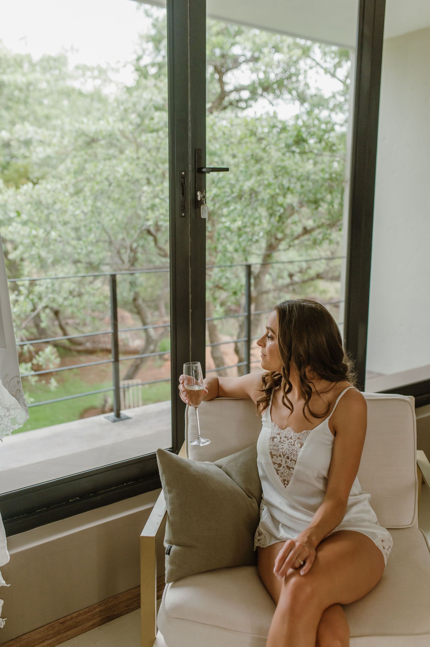 Woman in white lounges by large window with champagne glass overlooking garden view.