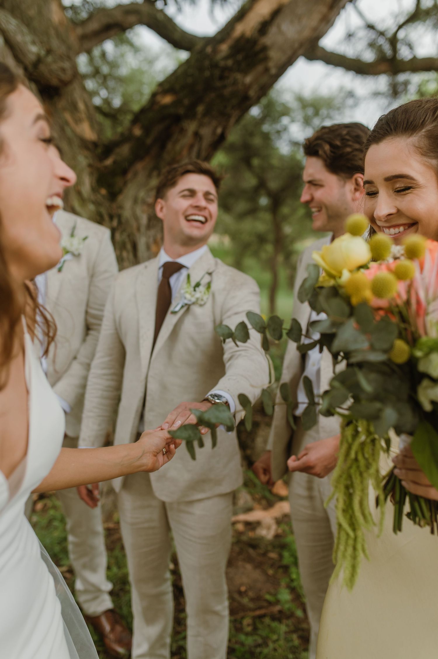 Wedding party members share joyful moments together under a tree with yellow floral accents.