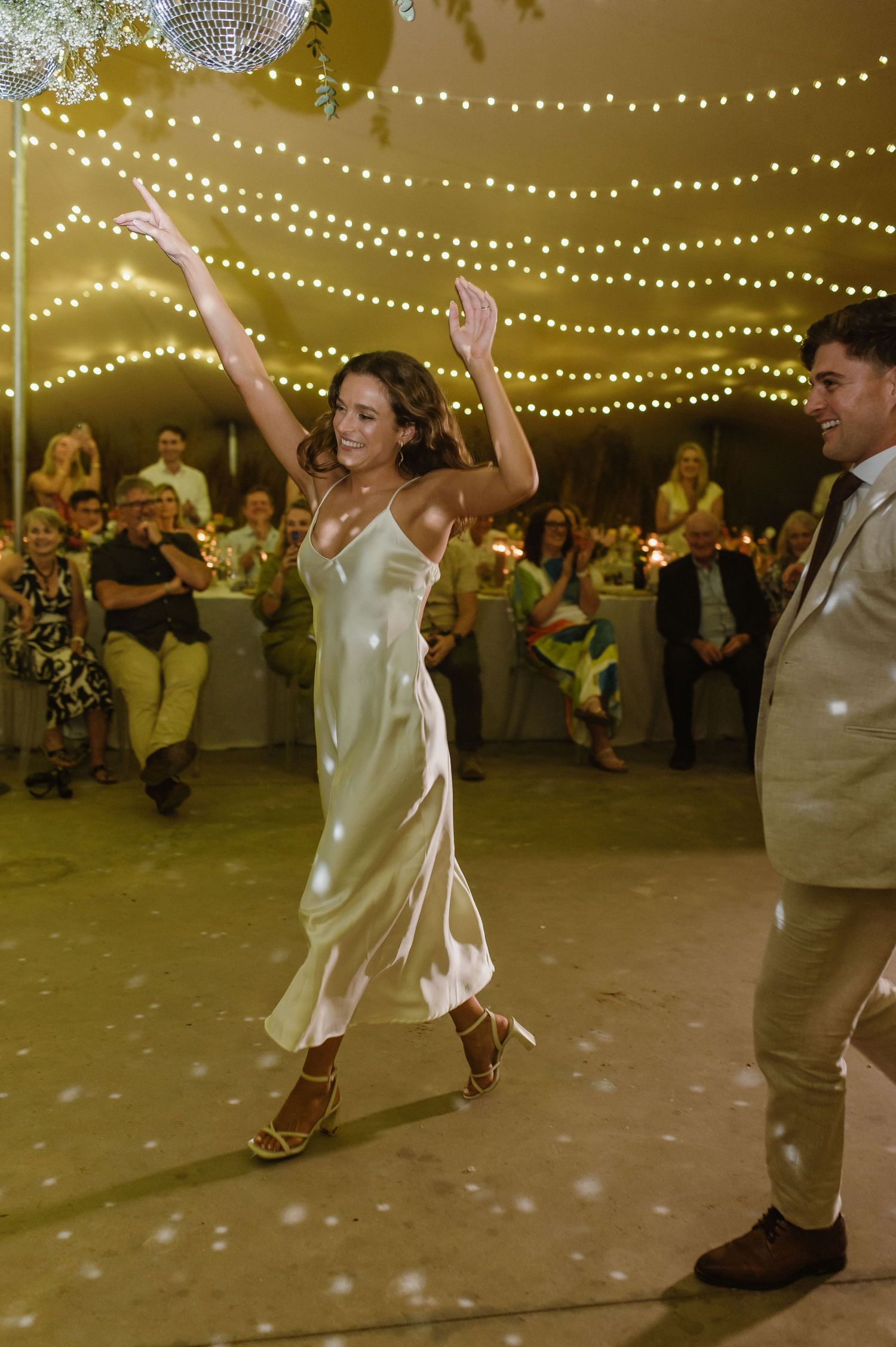 Wedding celebration dancers strike energetic poses under decorative string lights.