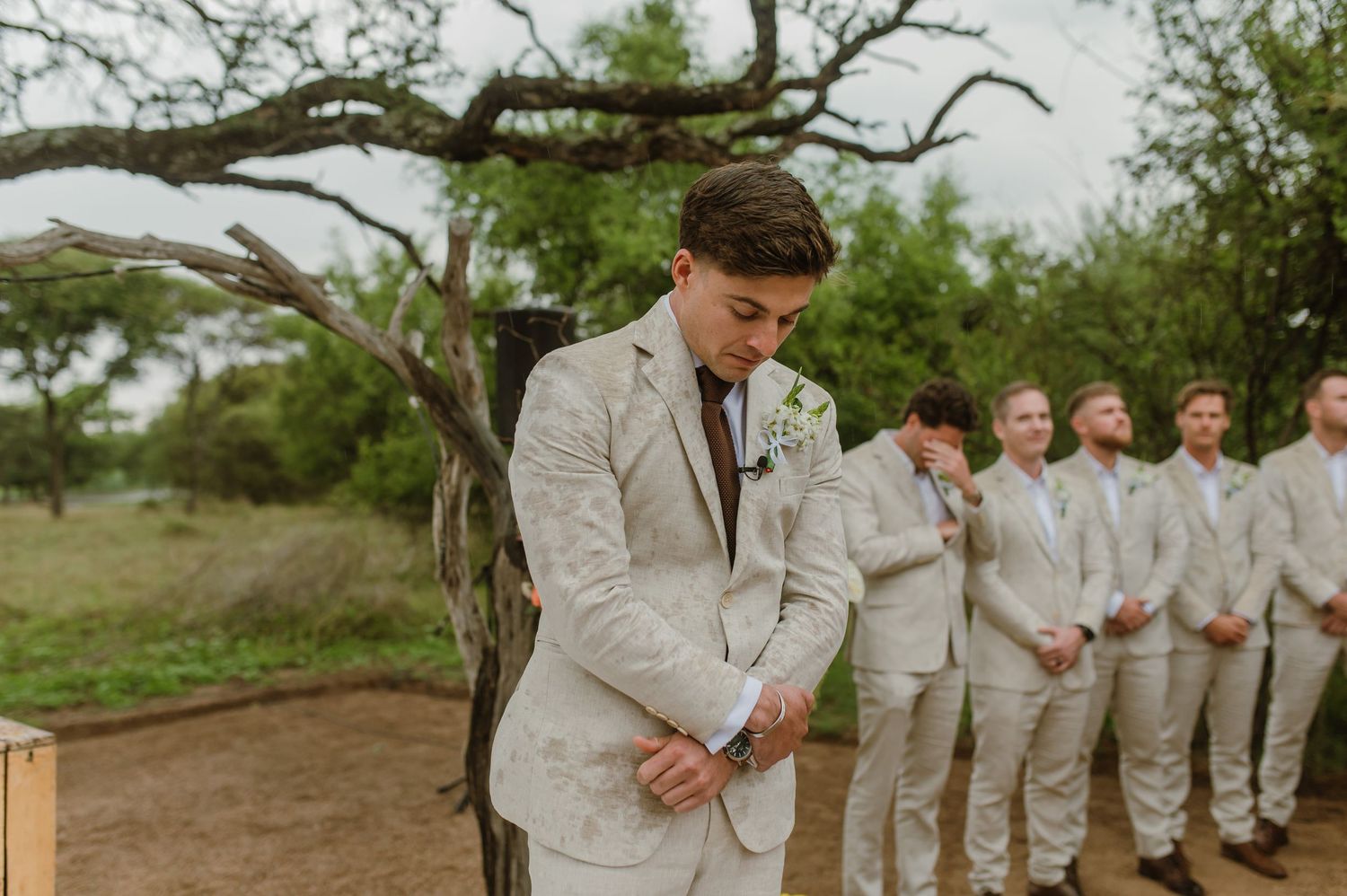 Wedding party members in light colored attire gather under sprawling tree branches.