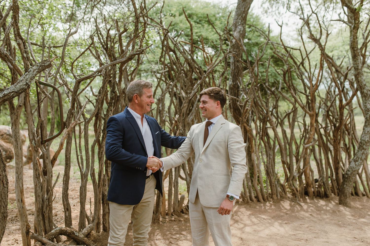 Groomsmen in light colored suits stand together in a wooded area for wedding photos.
