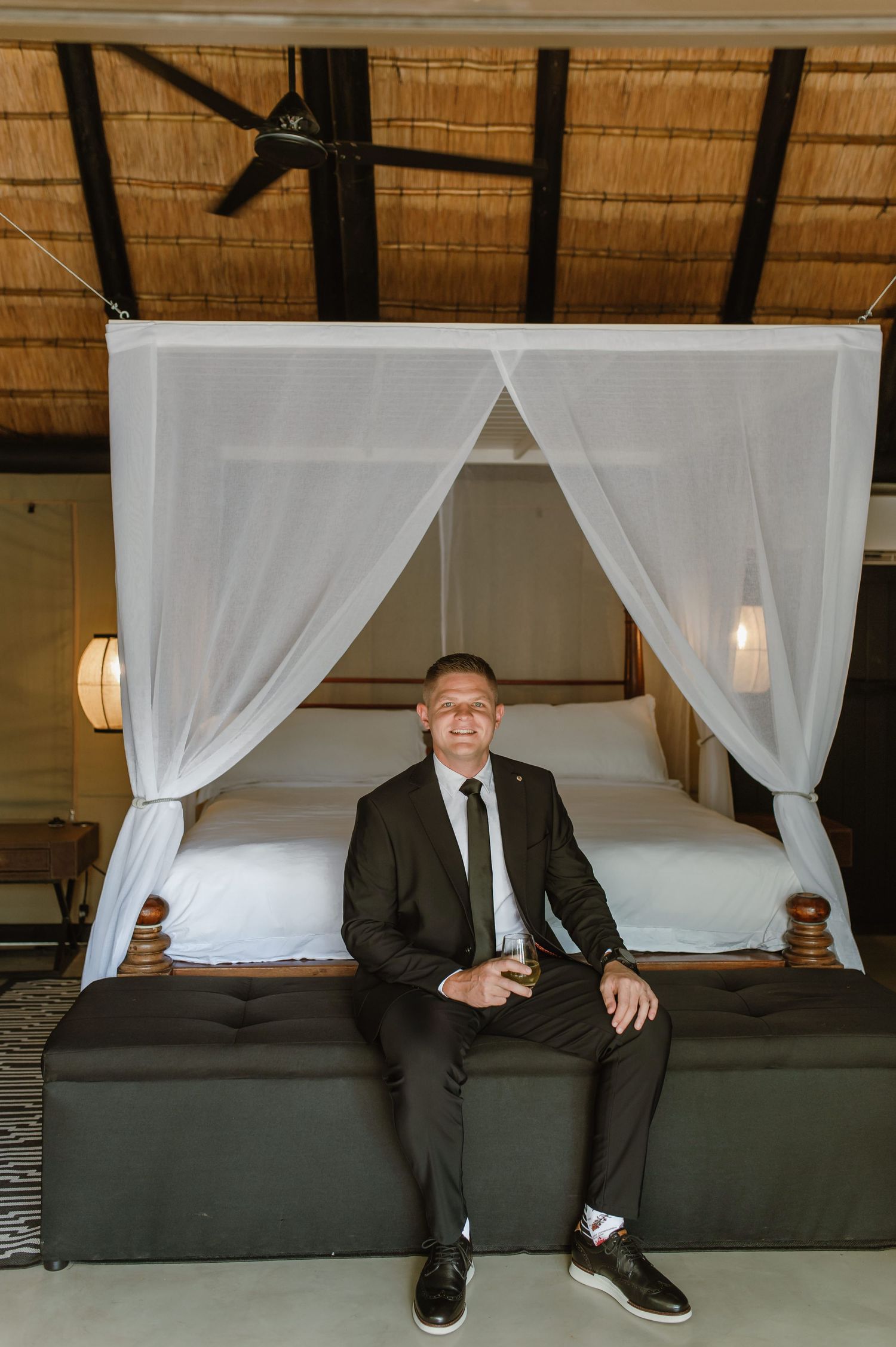 Groom in black suit sitting on canopy bed in safari lodge accommodation.