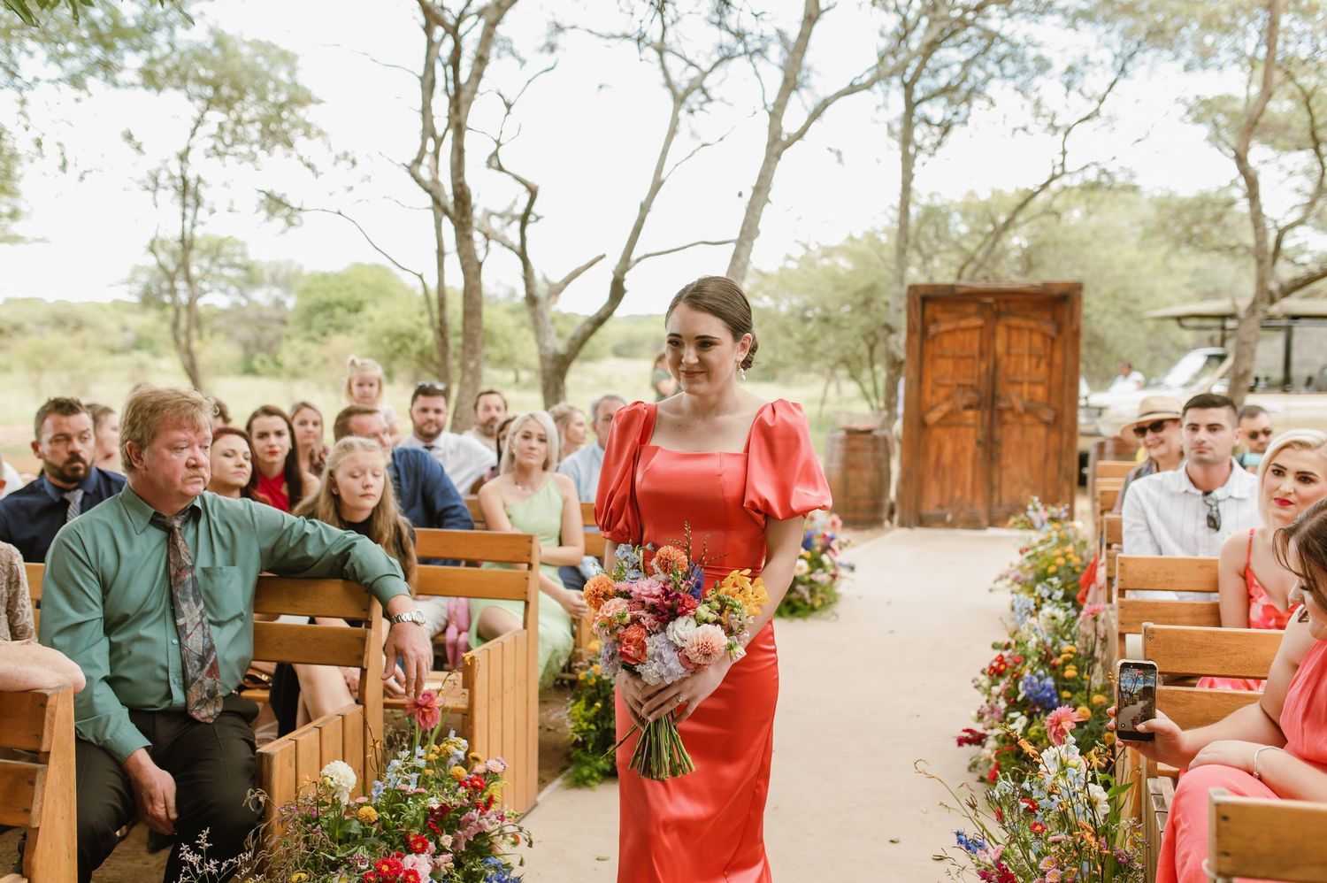 Bridesmaid in red dress walks down outdoor aisle carrying colorful bouquet at garden wedding.