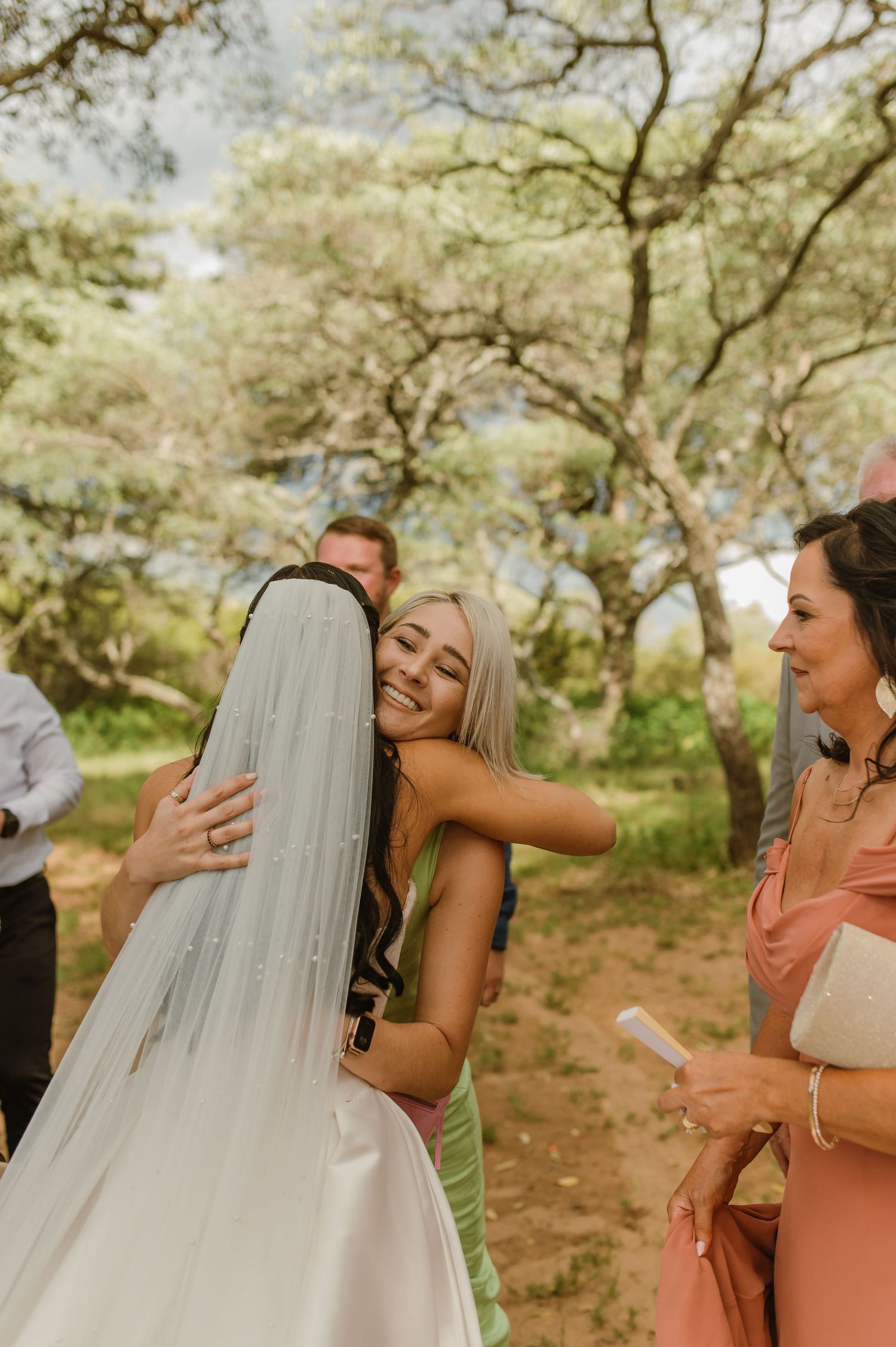 Blonde bridesmaid in green dress hugging bride in white veil at outdoor wedding celebration.
