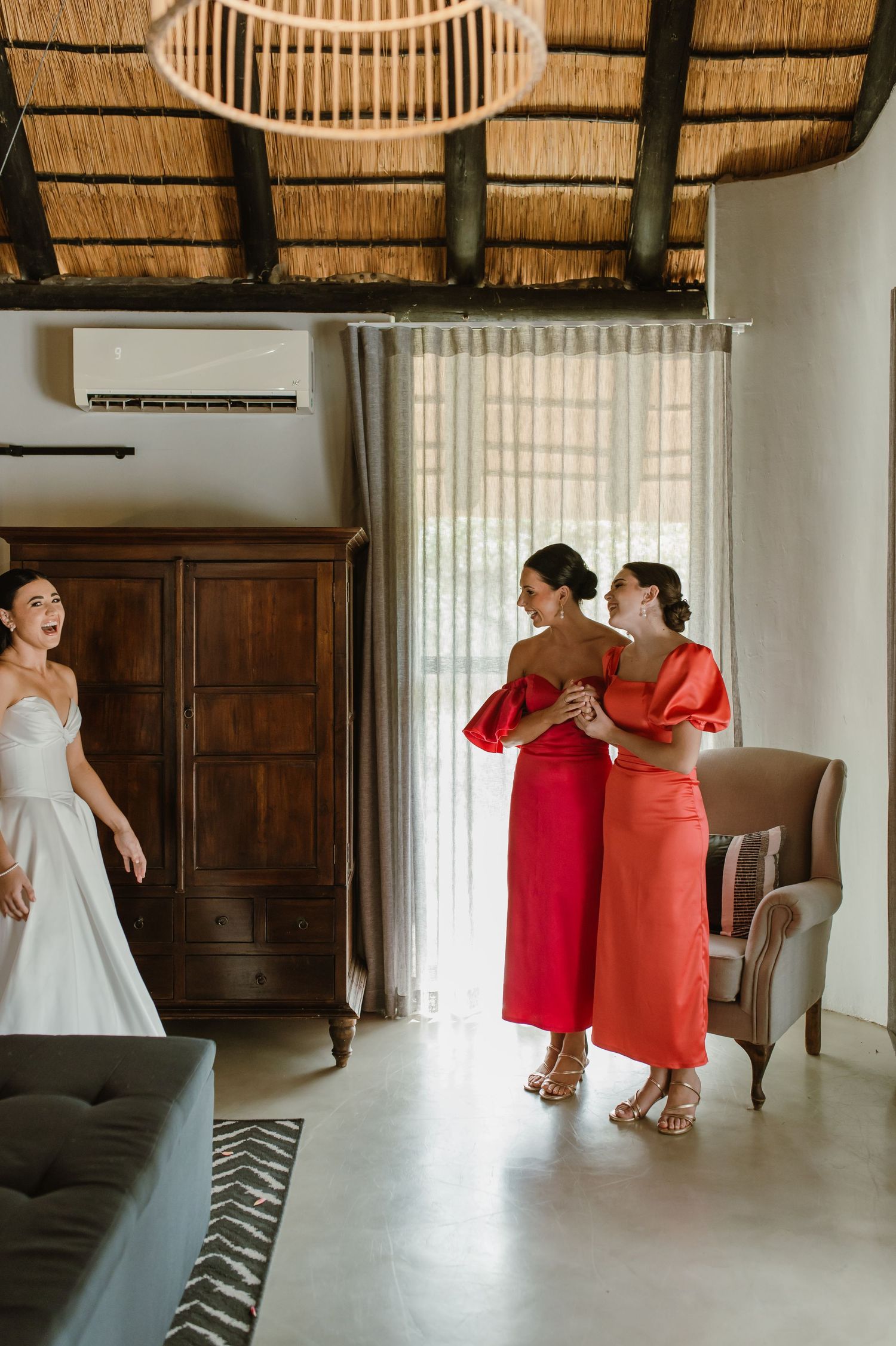 Bride in strapless gown surprises two bridesmaids in red and coral dresses indoors.