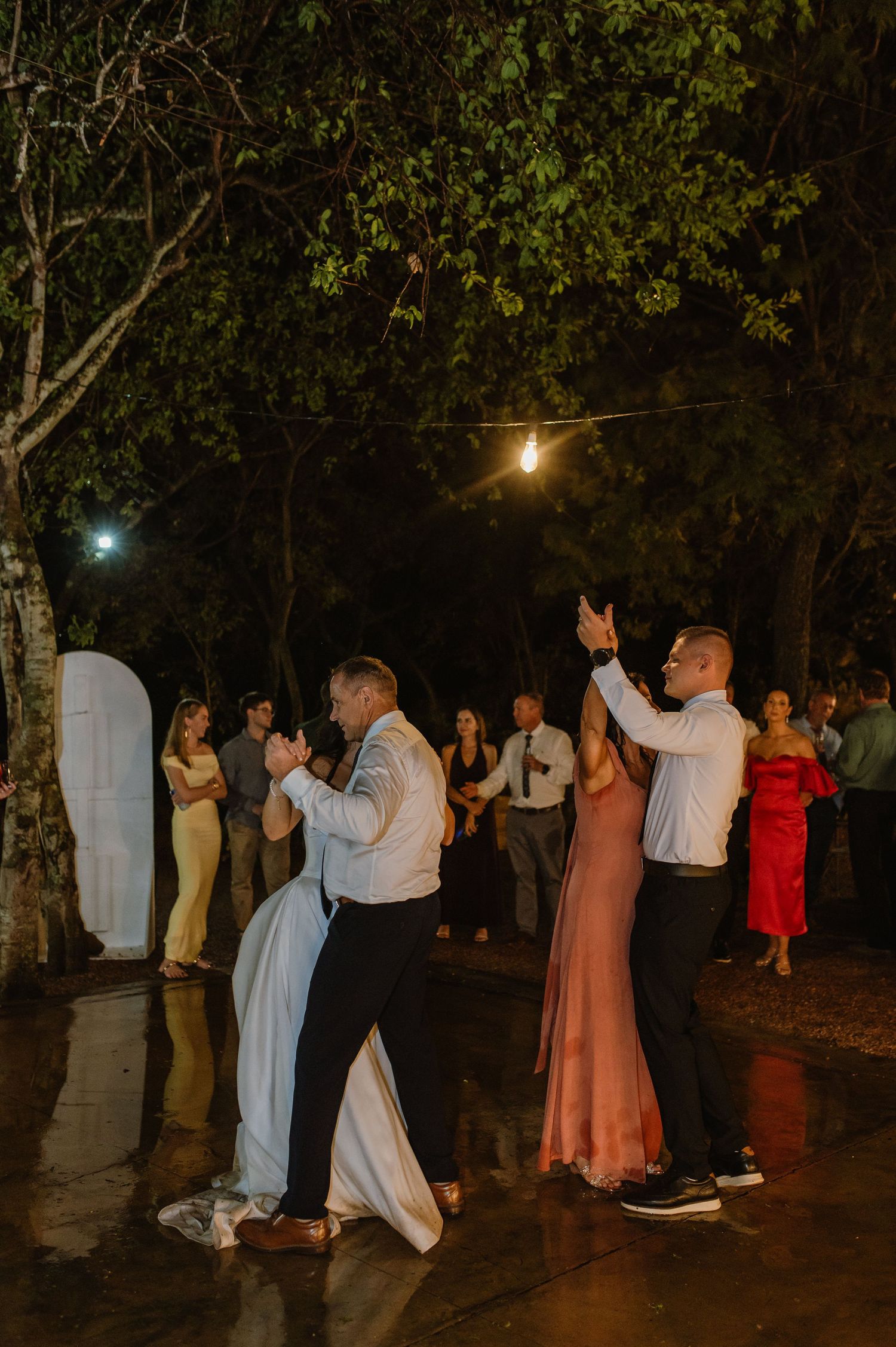 Wedding guests dancing outdoors at night under string lights surrounded by trees.