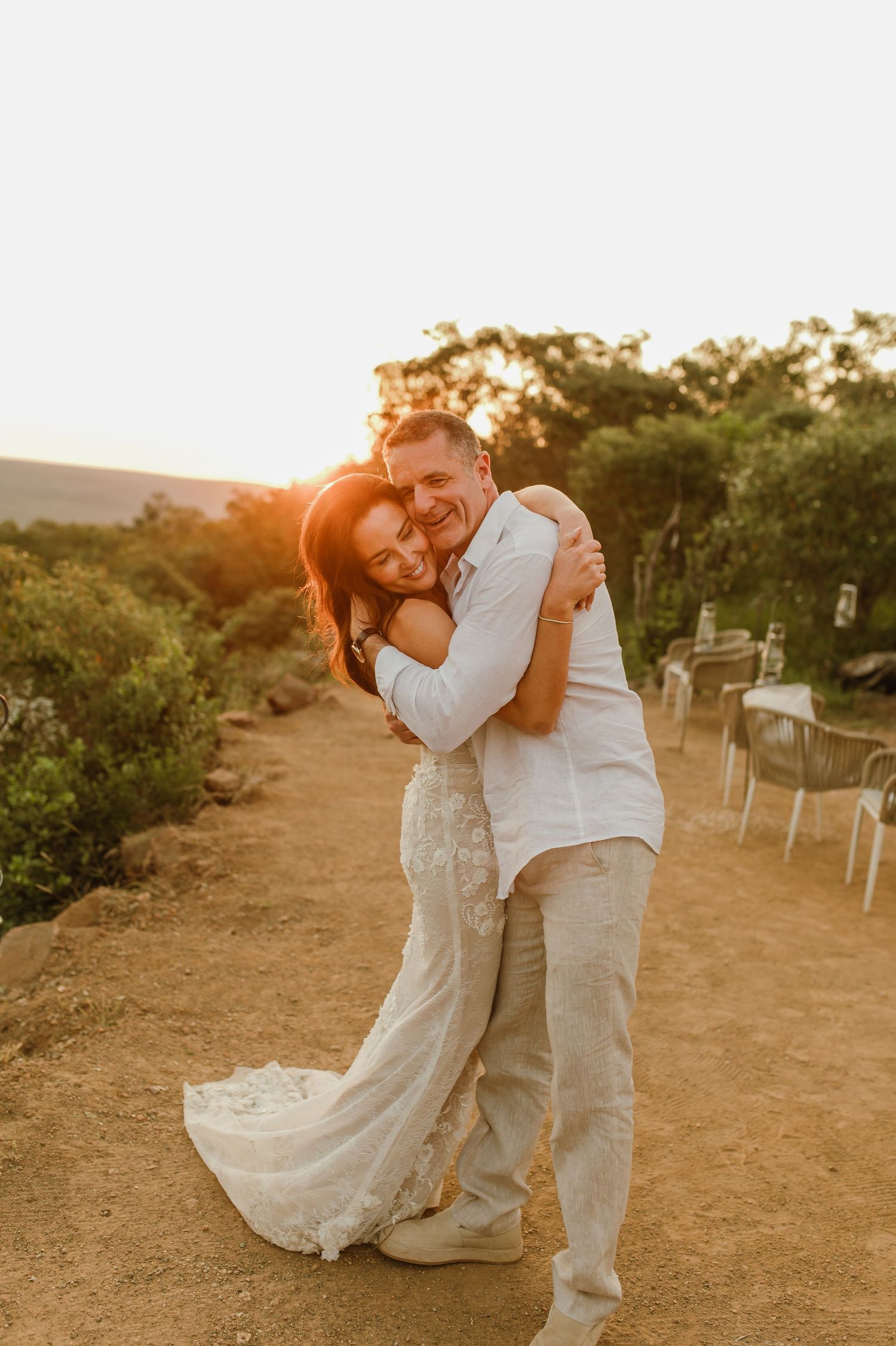 Joyful bride in lace gown hugs groom in white shirt during warm golden sunset outdoor wedding.