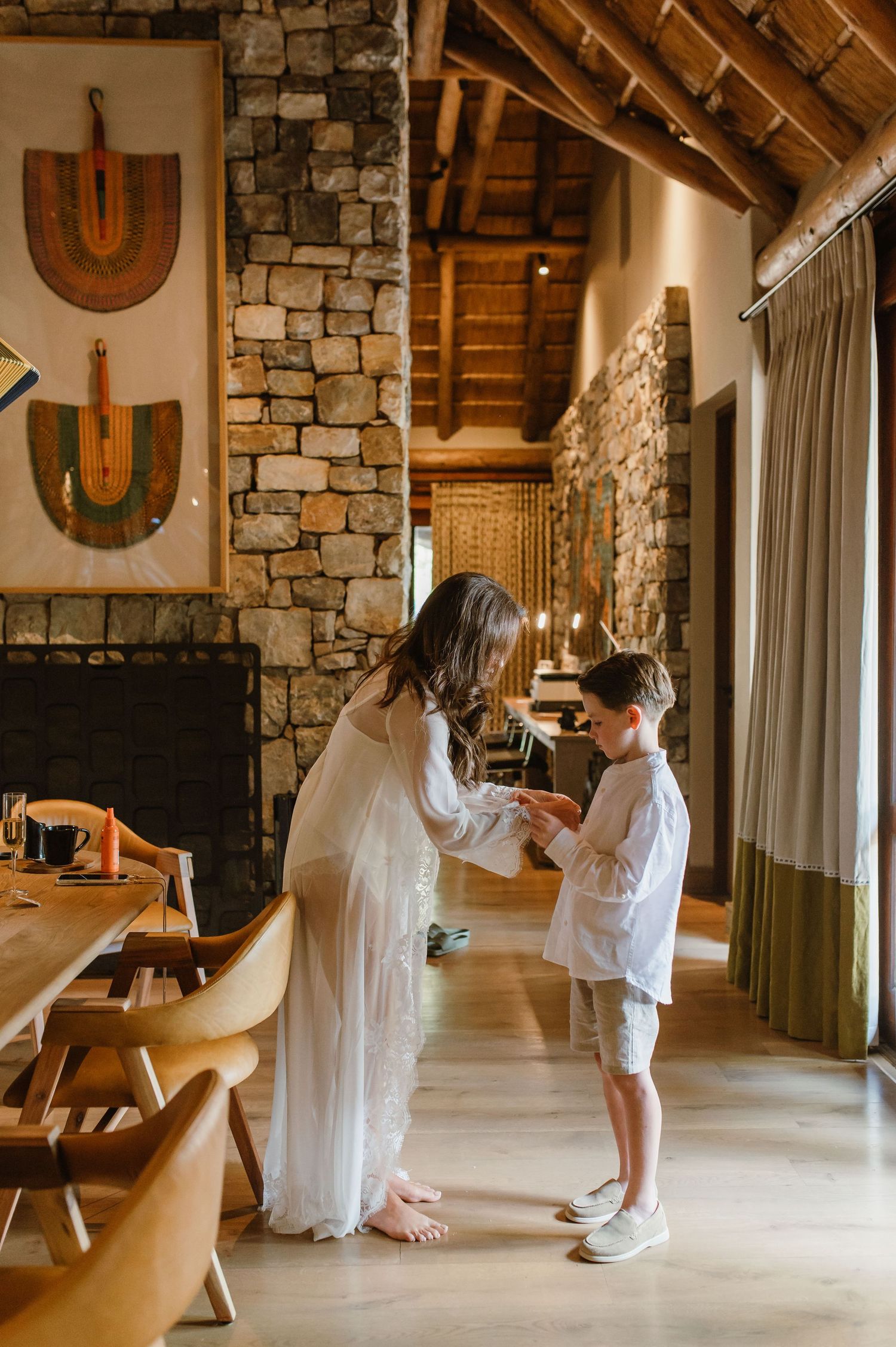 Woman in white dress adjusting a boy's shirt in a rustic stone lodge dining room interior.
