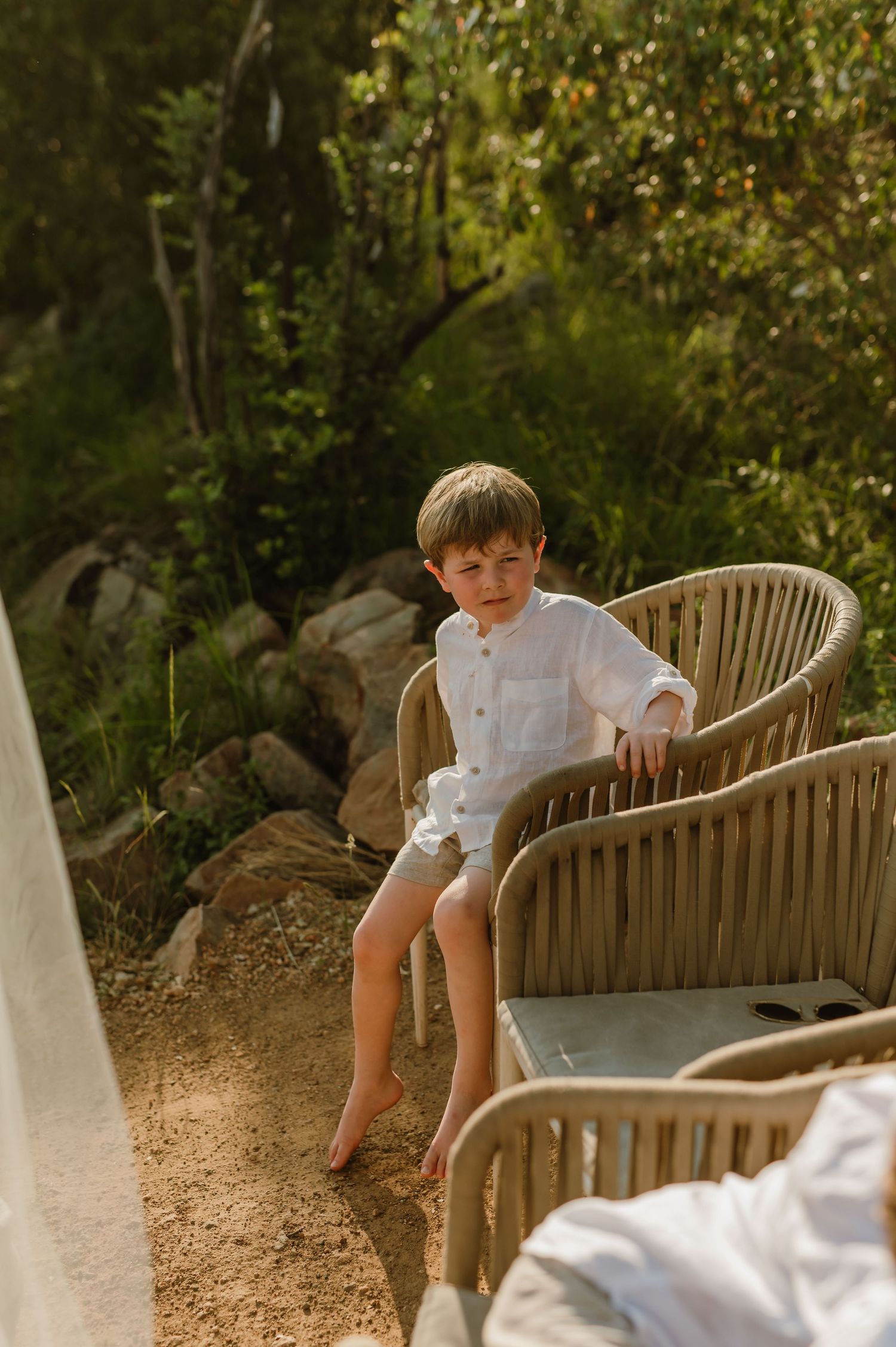 Young boy in white linen shirt and shorts sitting on wicker outdoor chair in garden setting.
