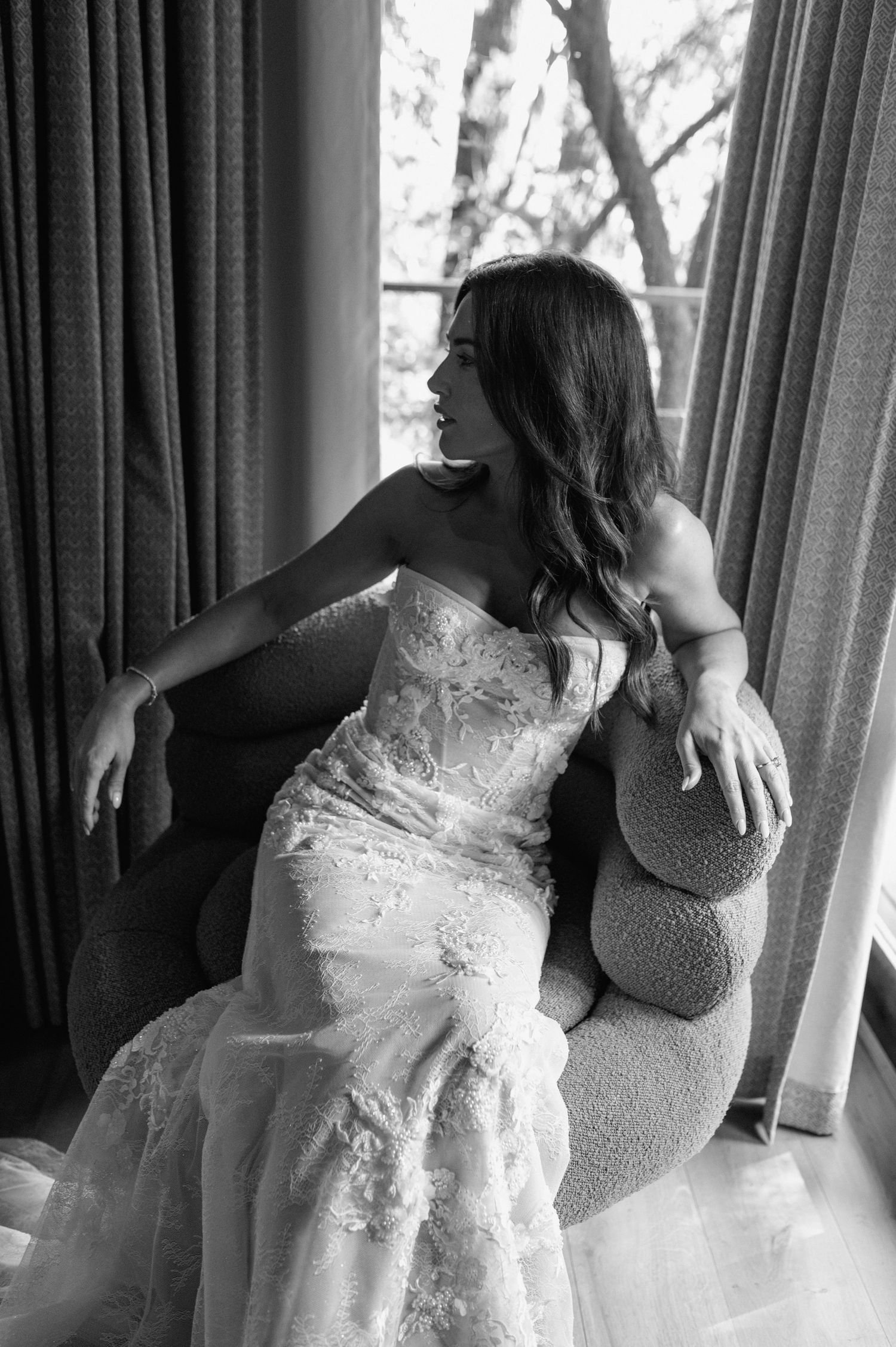 Black and white photo of bride in strapless lace wedding gown seated in chair by window.