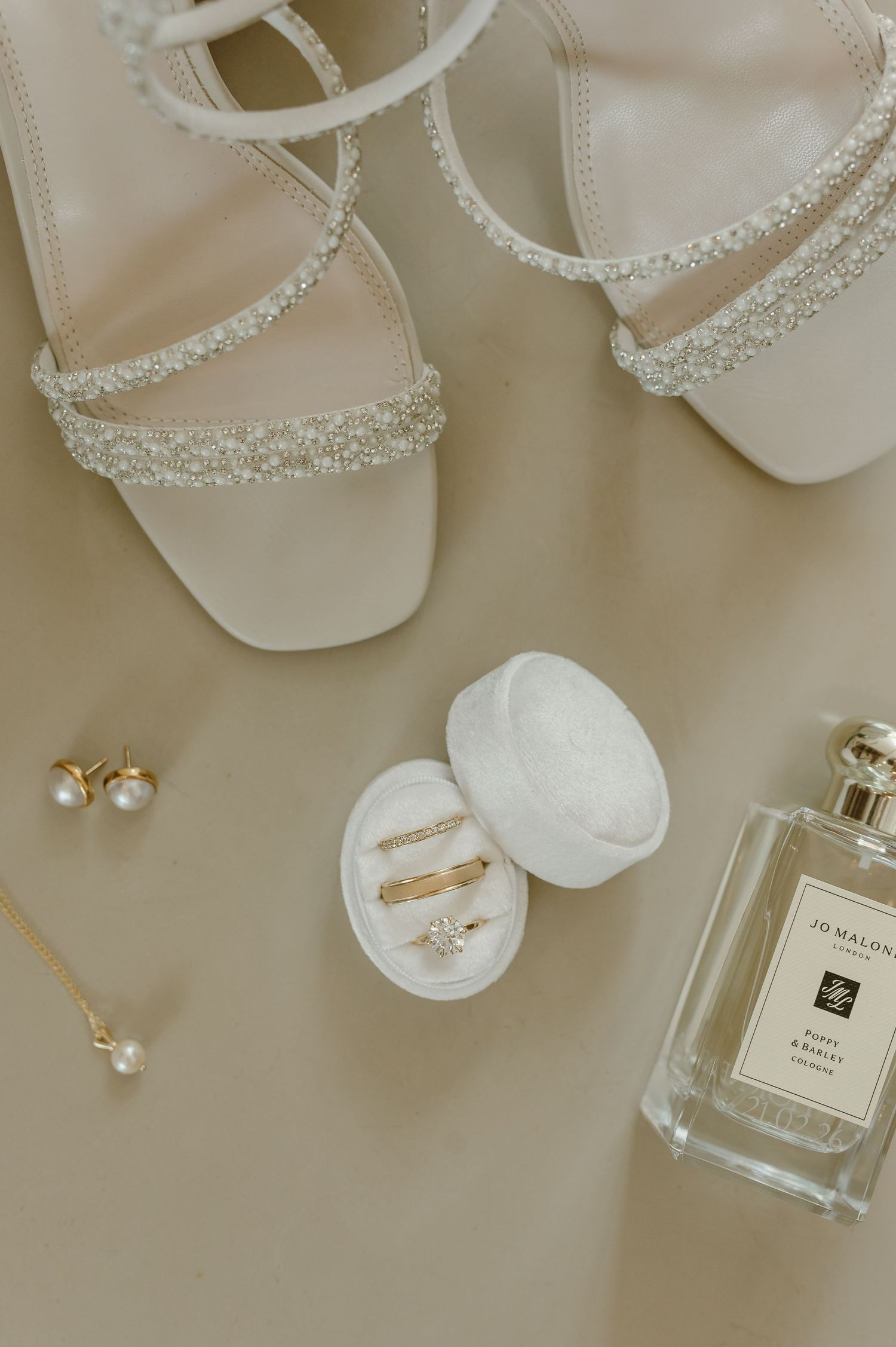 Wedding accessories including pearl-trimmed shoes, jewelry box, and Jo Malone perfume bottle on beige surface.