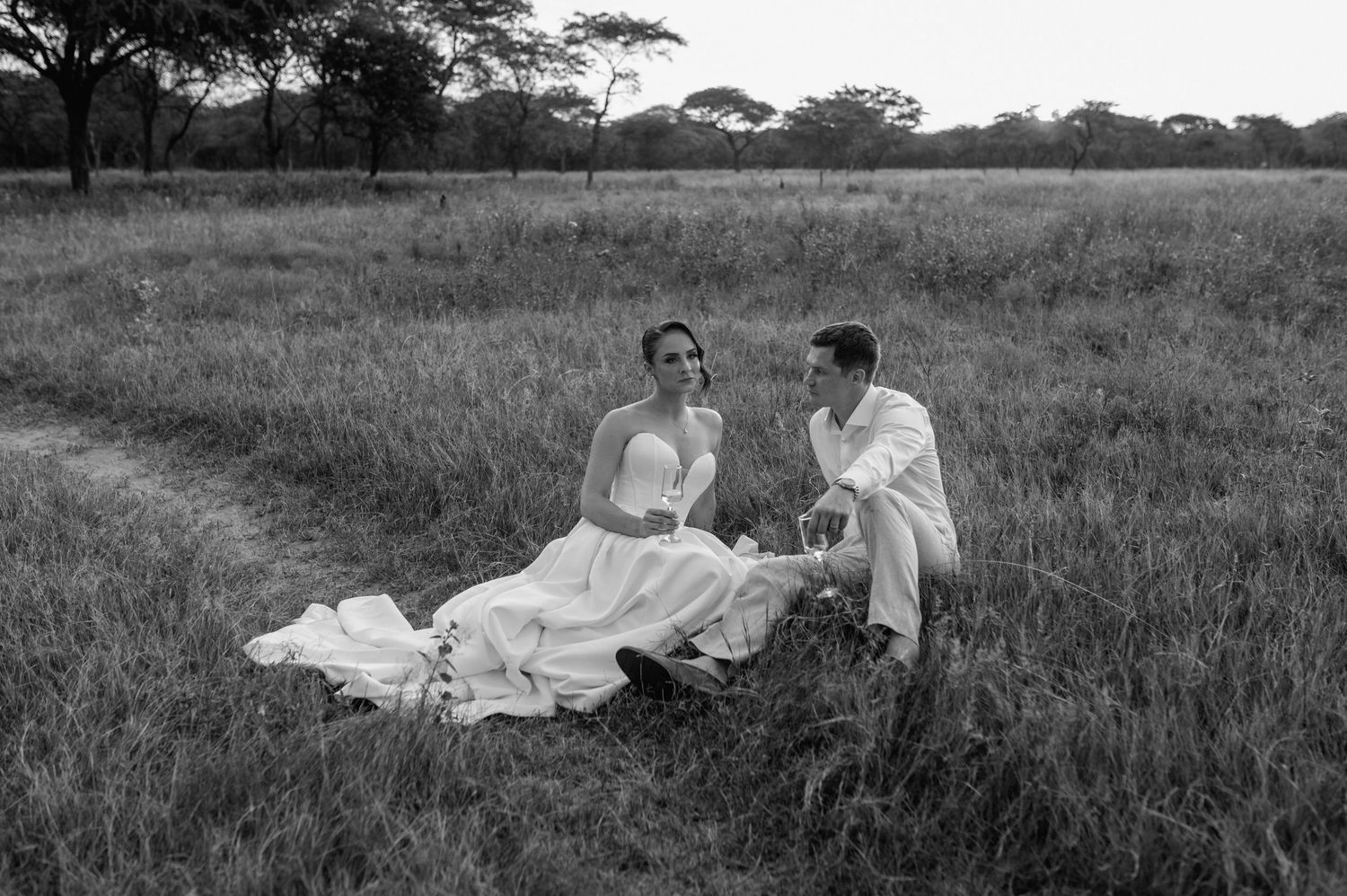 A couple in wedding attire sits on a blanket in a grassy field during a black and white outdoor photoshoot.