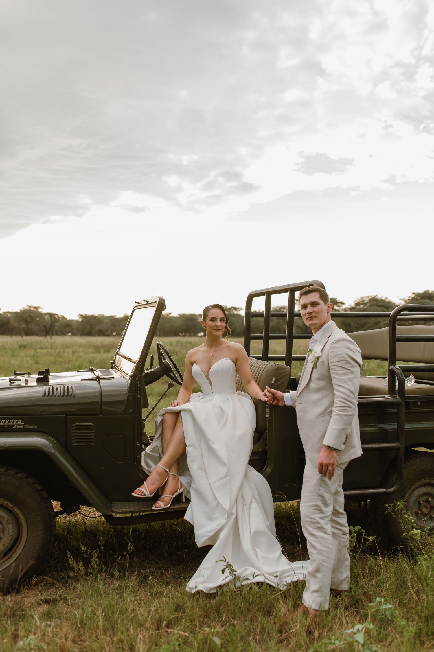 A couple in wedding attire poses playfully by a vintage black car in a rustic field setting.