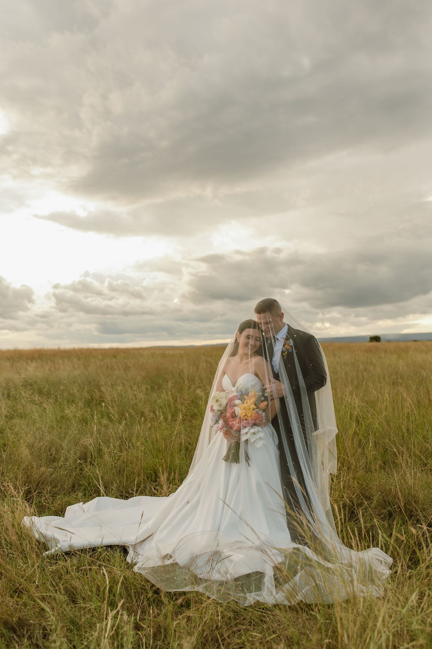 Bride and groom embracing in golden grassland with dramatic cloudy sky backdrop.