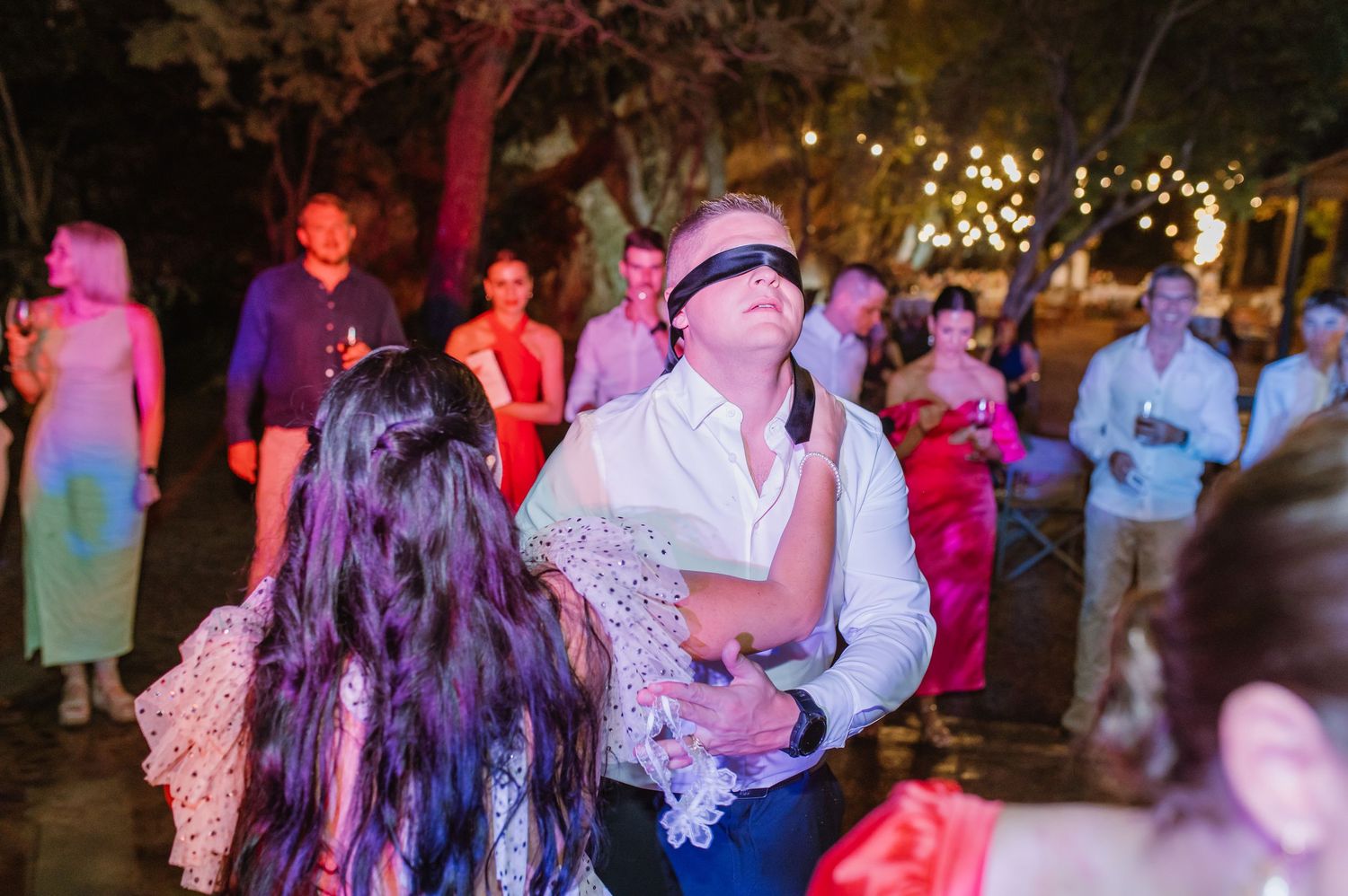 Blindfolded groom dances with bride at lively outdoor wedding reception under string lights at night.