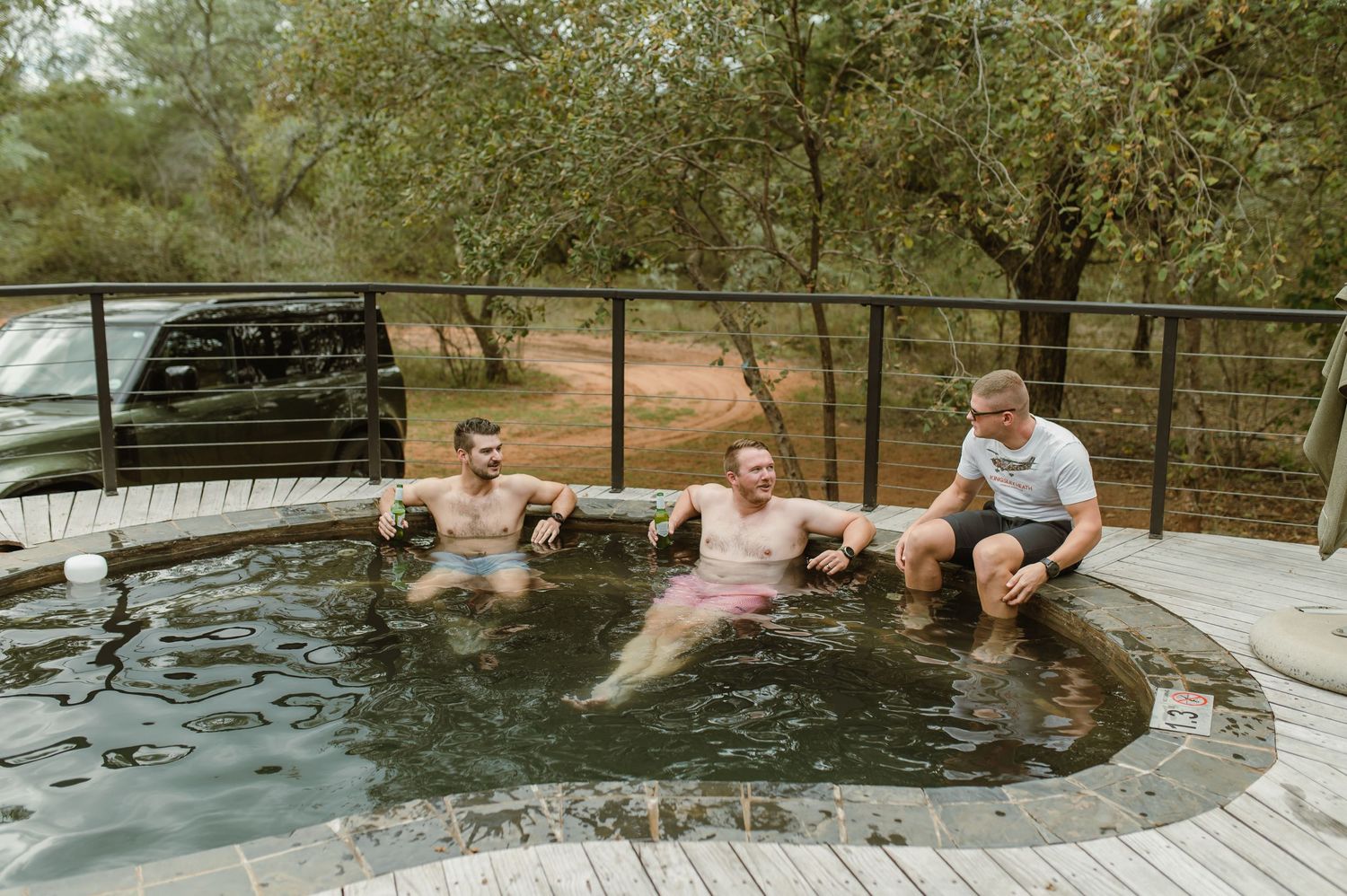 Three men relaxing in an outdoor hot tub surrounded by trees and a wooden deck in a natural setting.