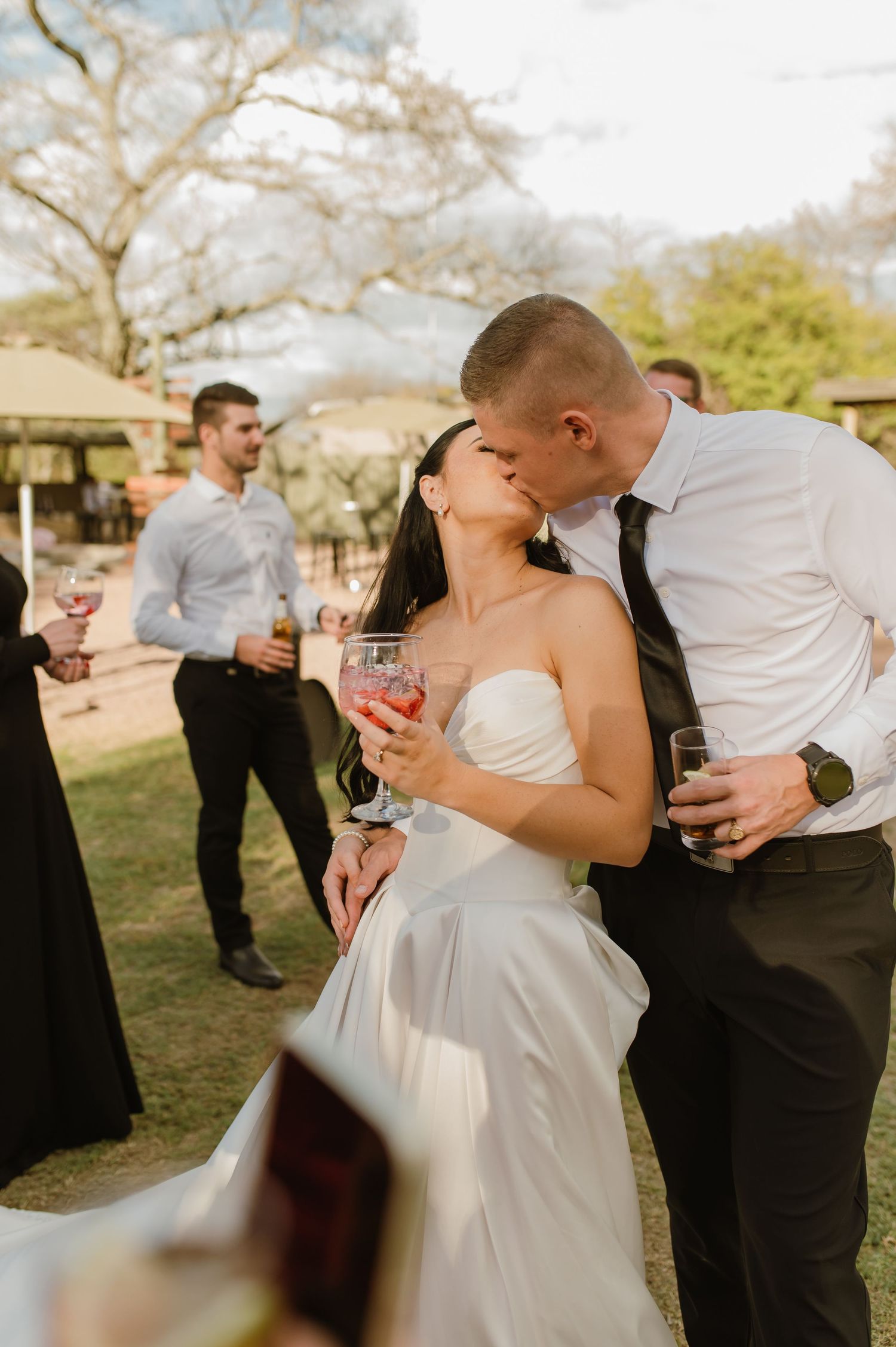 Newlyweds sharing a kiss while holding drinks at their outdoor wedding reception.